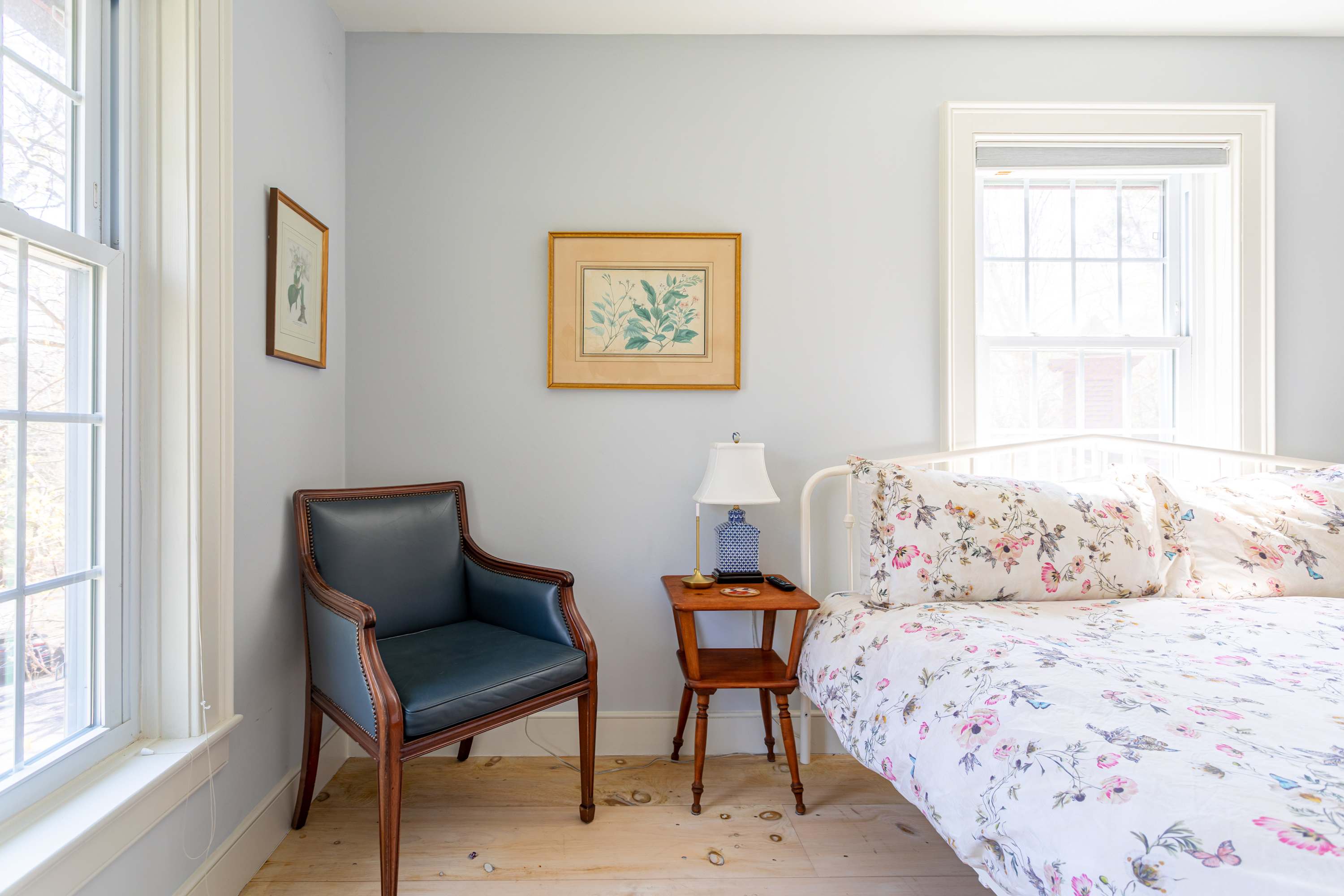 Light-filled bedroom with pale blue walls, light wood floors, a white metal bed frame with floral bedding, a dark wood armchair, and a small wooden side table with a lamp. Two framed botanical prints adorn the wall.