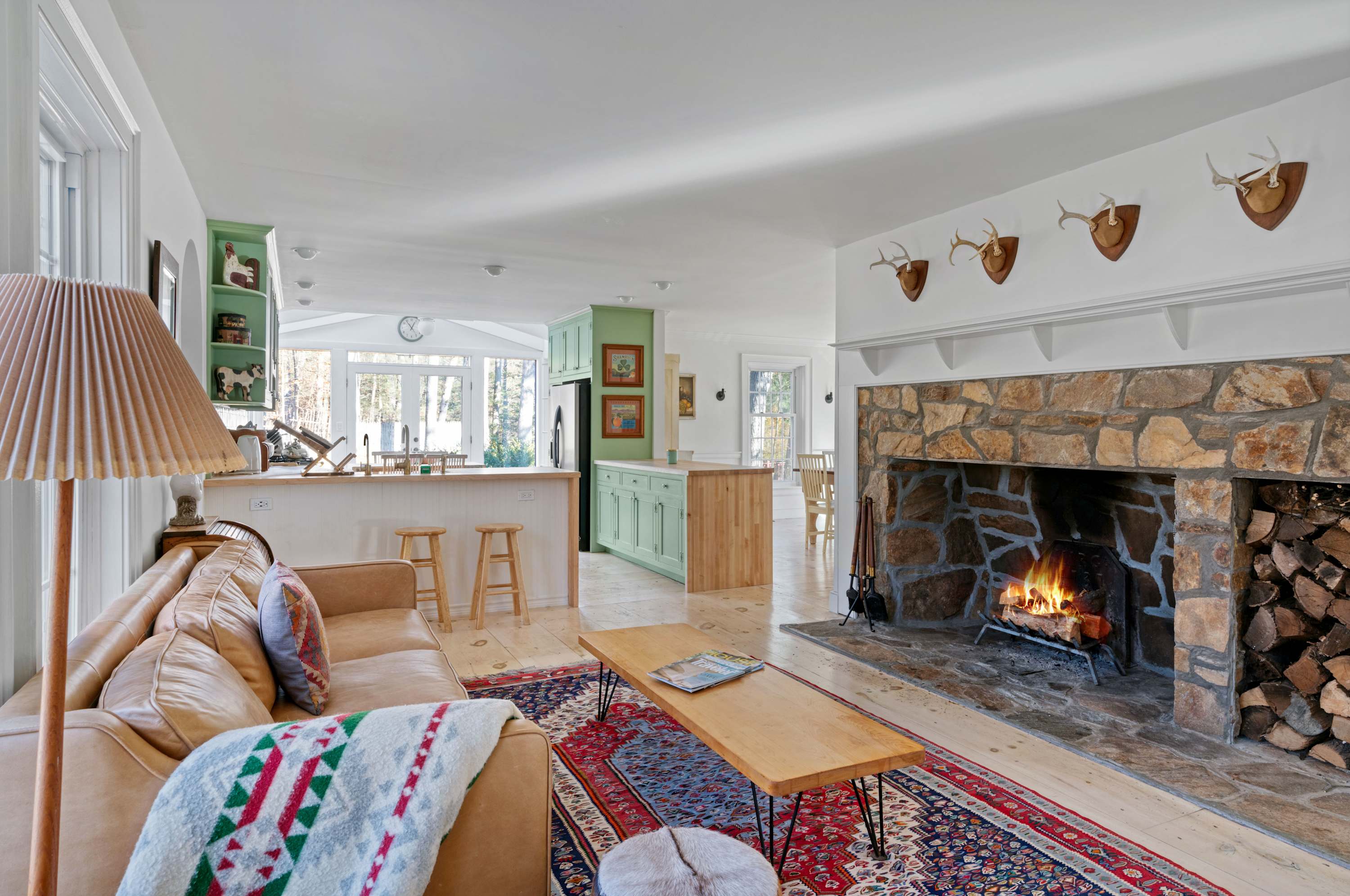 A spacious living room featuring a large tan leather sofa with colorful throw pillows and a wooden coffee table on a vibrant patterned rug. A grand stone fireplace with a roaring fire and stacked firewood is a prominent focal point.