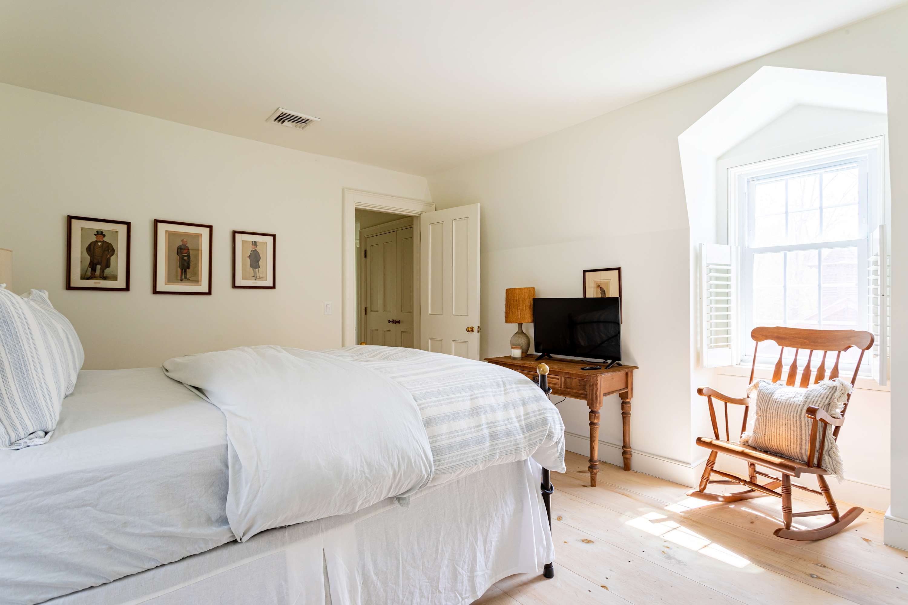 View of the bright bedroom with white walls and light wood floors, showing the black and brass bed, a wooden side table with a TV, and a wooden rocking chair by a window with white shutters. Three framed portraits hang above the bed.