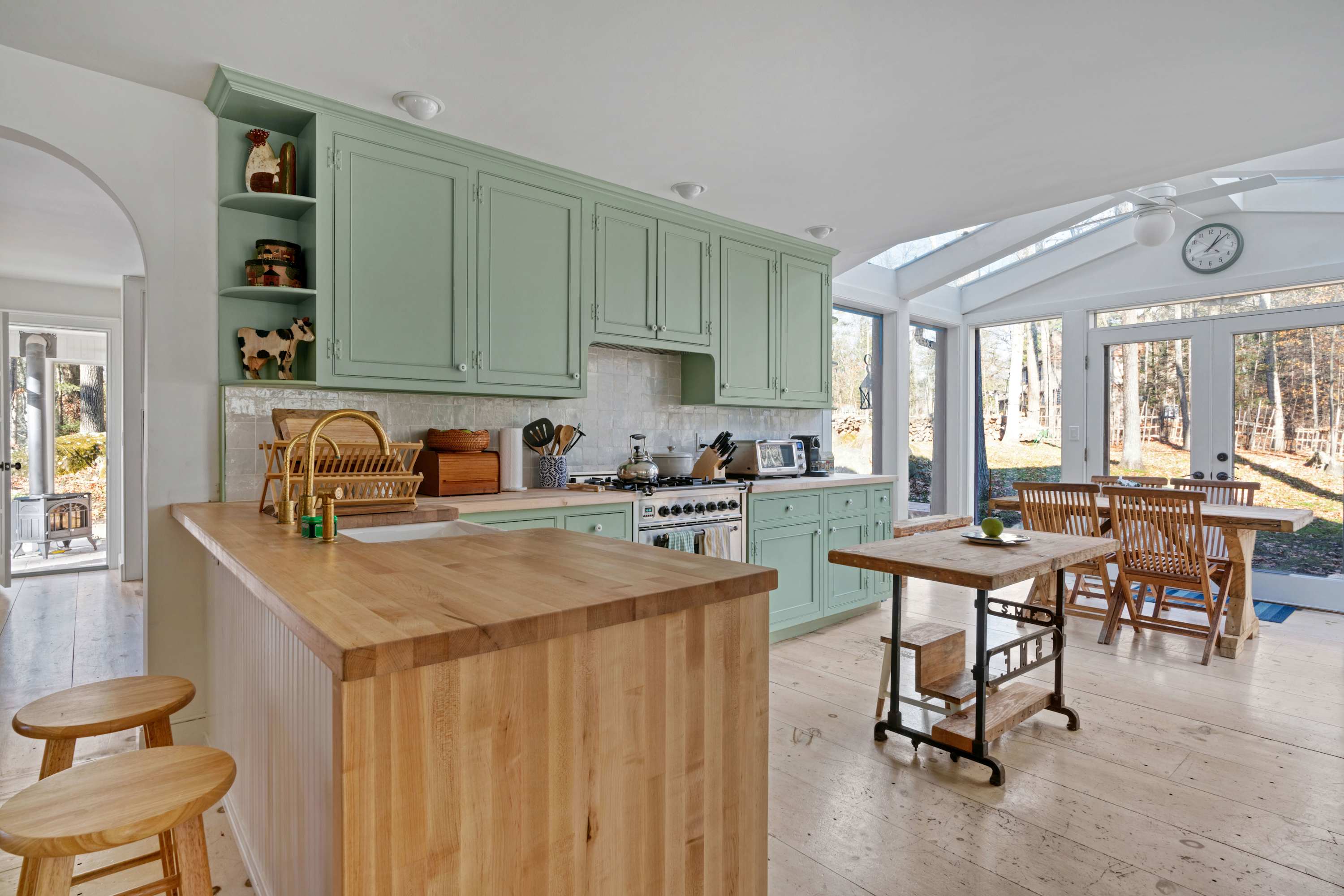 A bright kitchen featuring light green shaker-style cabinets, light wood butcher block countertops, and a stainless steel range. A wooden island with bar stools and a small dining table extend into a sunlit dining area with large windows.