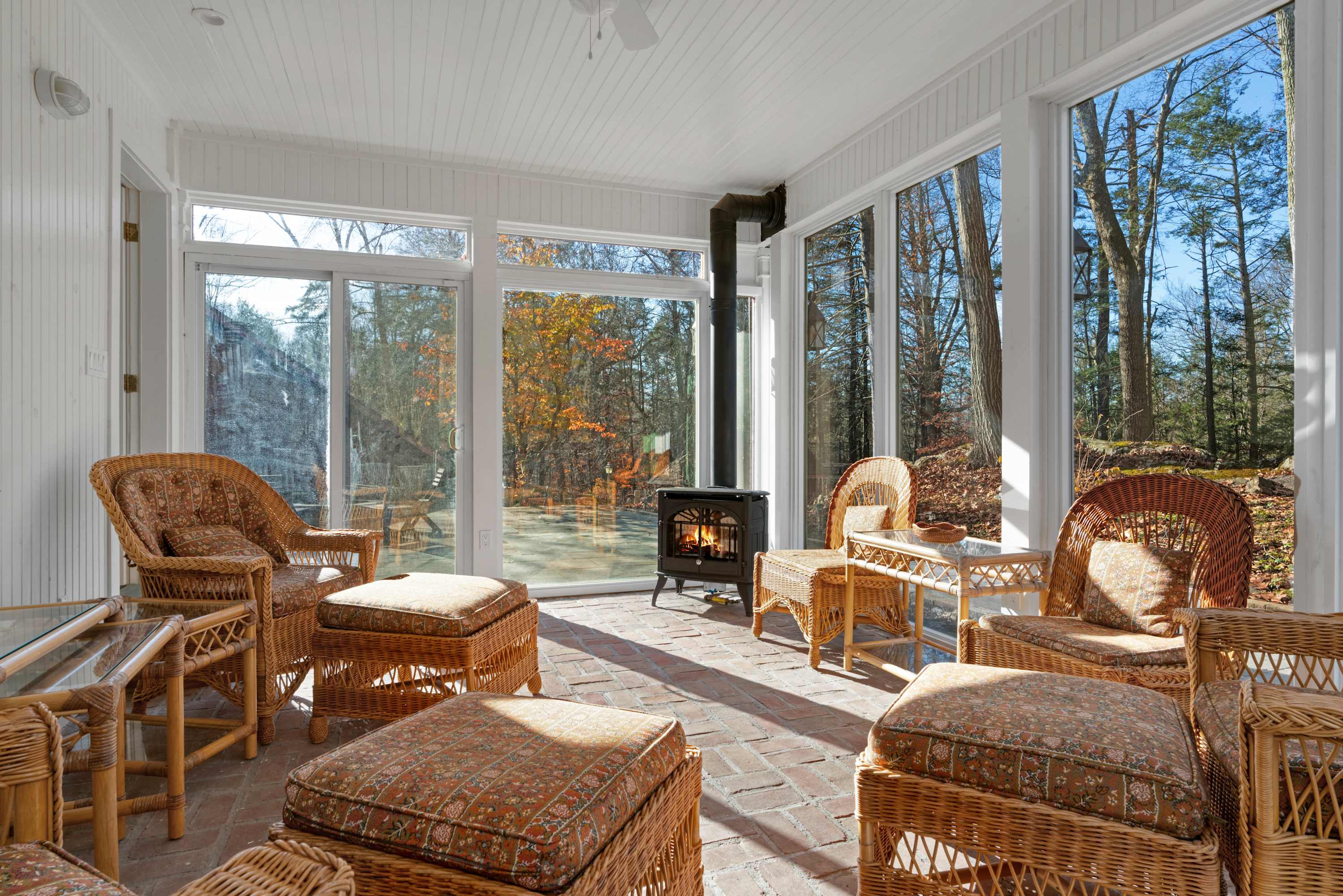 A bright sunroom features large windows offering panoramic views of the surrounding autumn woods, a cozy wood-burning stove, and a collection of wicker furniture with patterned cushions. The rustic brick floor adds warmth and character to the inviting space.