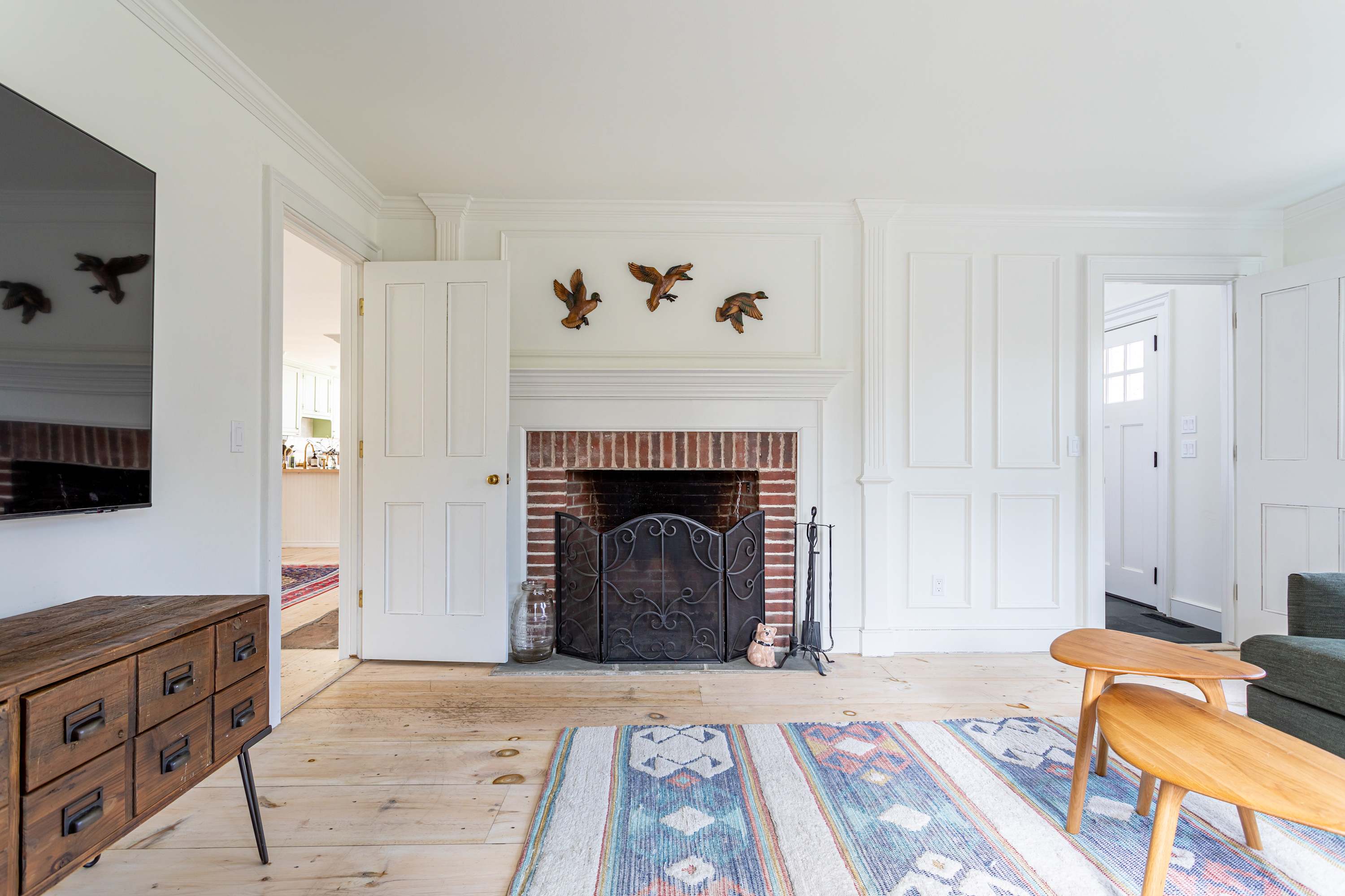 A traditional living room fireplace with a brick surround and a black metal screen, topped by a white wooden mantel adorned with carved wooden ducks. White paneled walls and light wide plank wood floors complete the classic look.