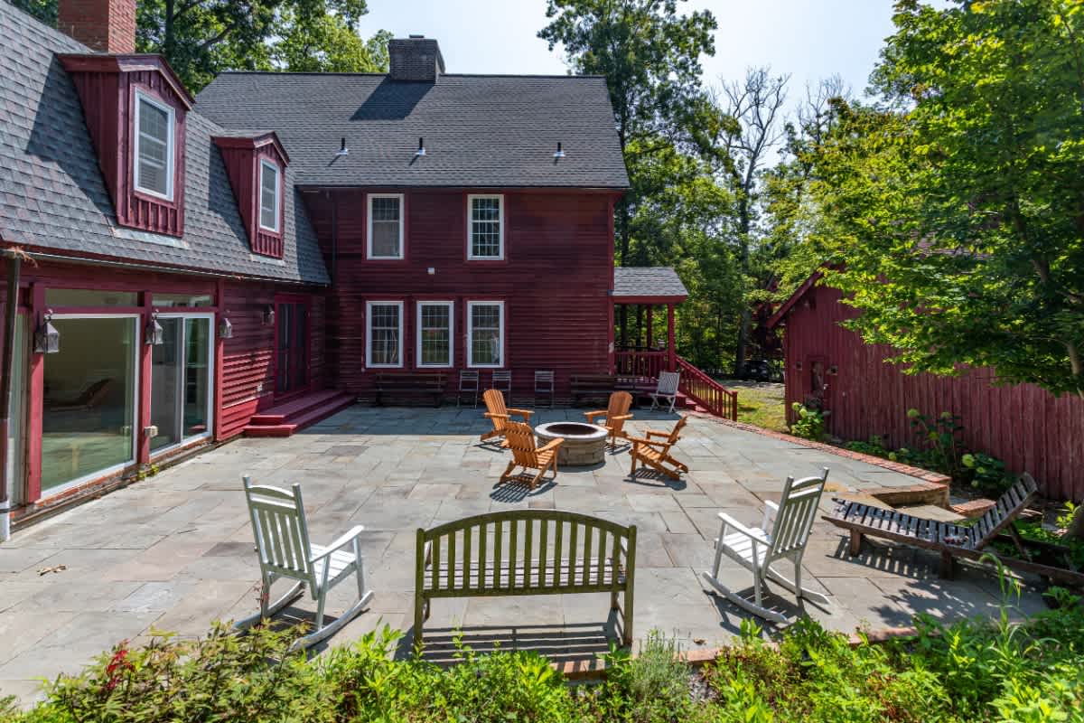 Spacious stone patio behind a red house, furnished with a fire pit, wooden Adirondack chairs, a bench, and white rocking chairs, surrounded by mature trees and a smaller red outbuilding.