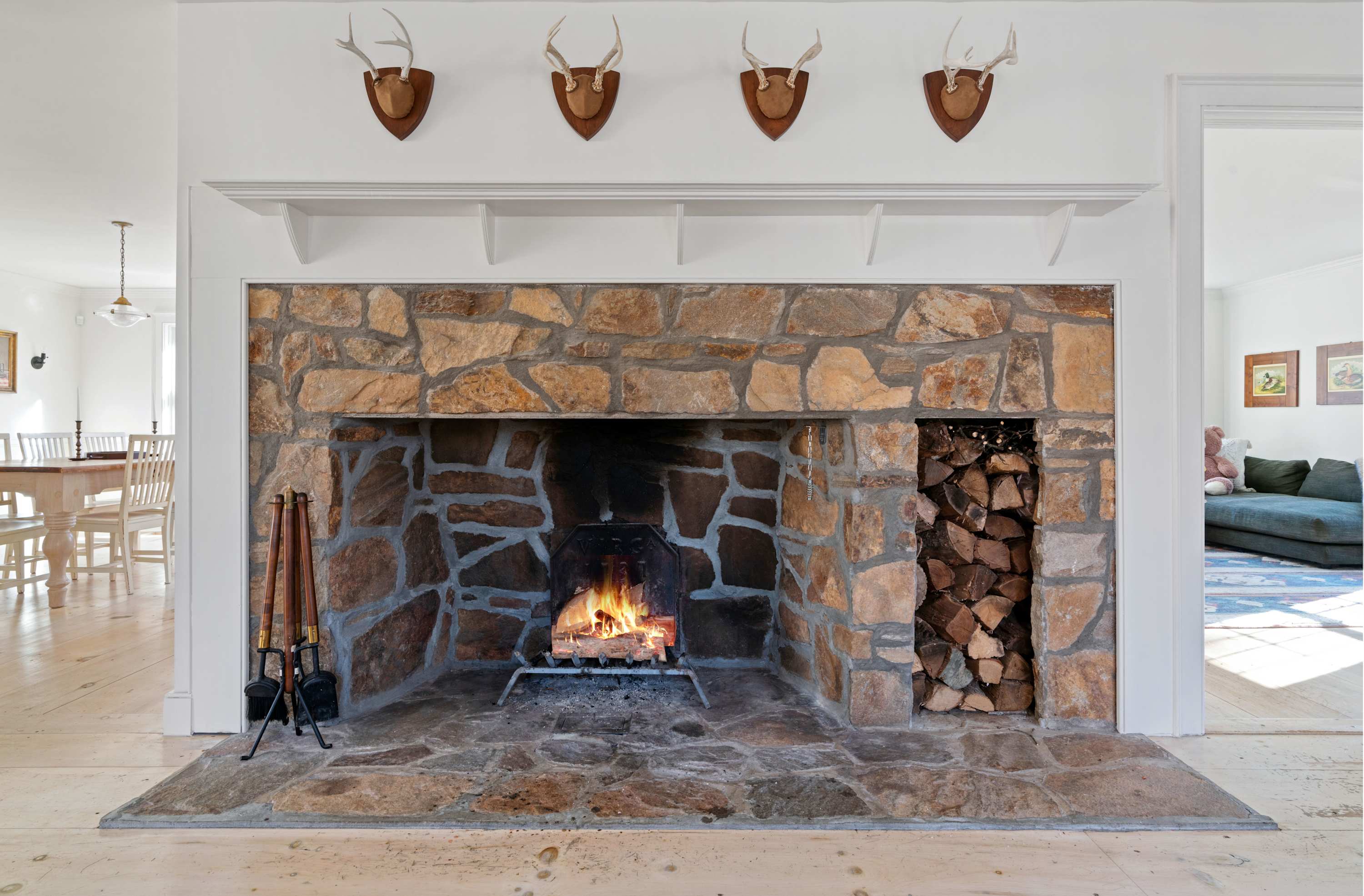 A grand stone fireplace with a burning fire and an integrated wood storage nook serves as the focal point of this living space, featuring light wood floors and decorative antler mounts above the white mantel. An open doorway provides a glimpse into an adjacent dining area.