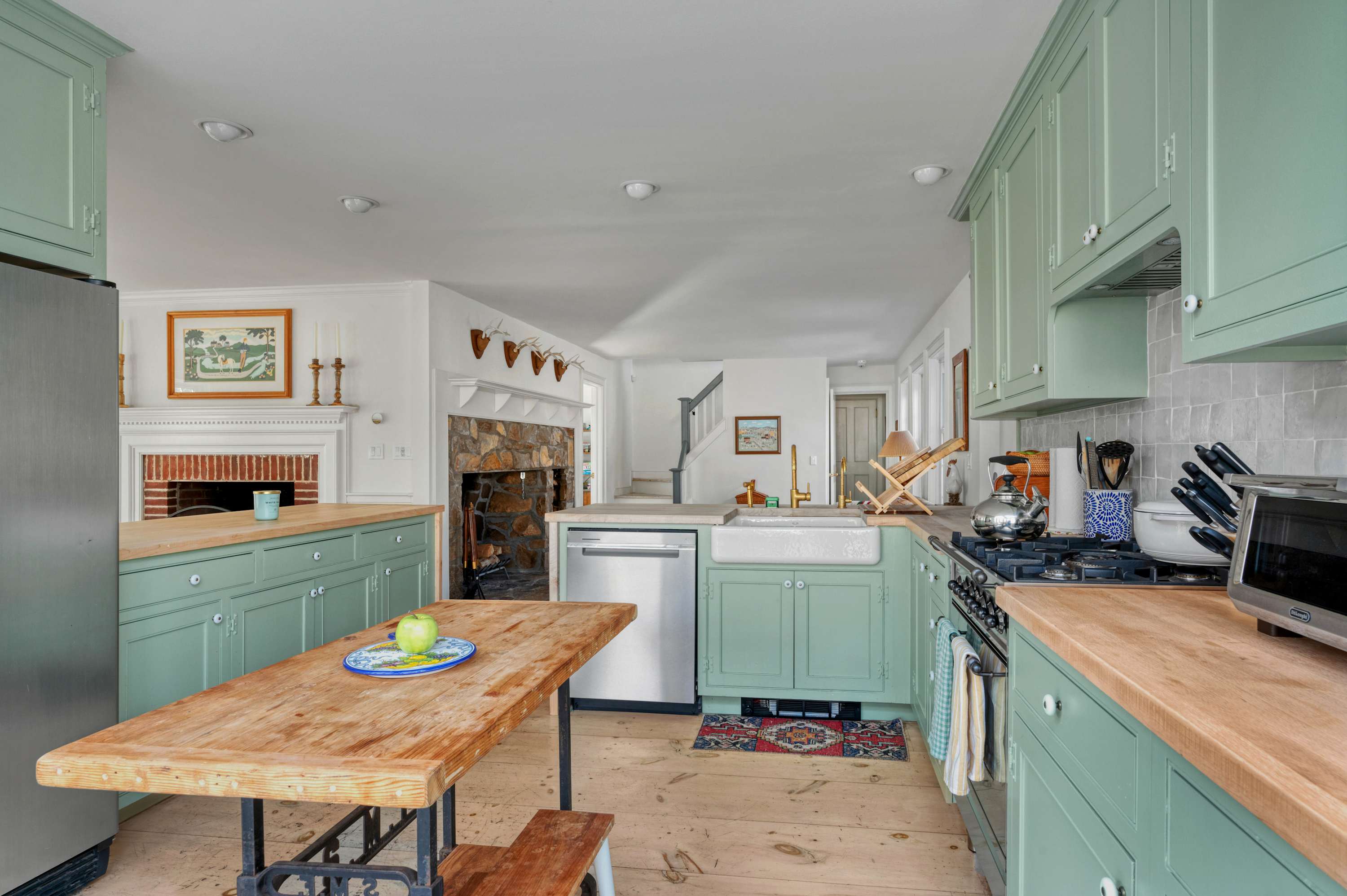 A spacious kitchen with light green cabinets, butcher block countertops, and a stainless steel refrigerator and dishwasher. A rustic wooden table with a green apple sits in the foreground, and the living room fireplace is visible in the distance.