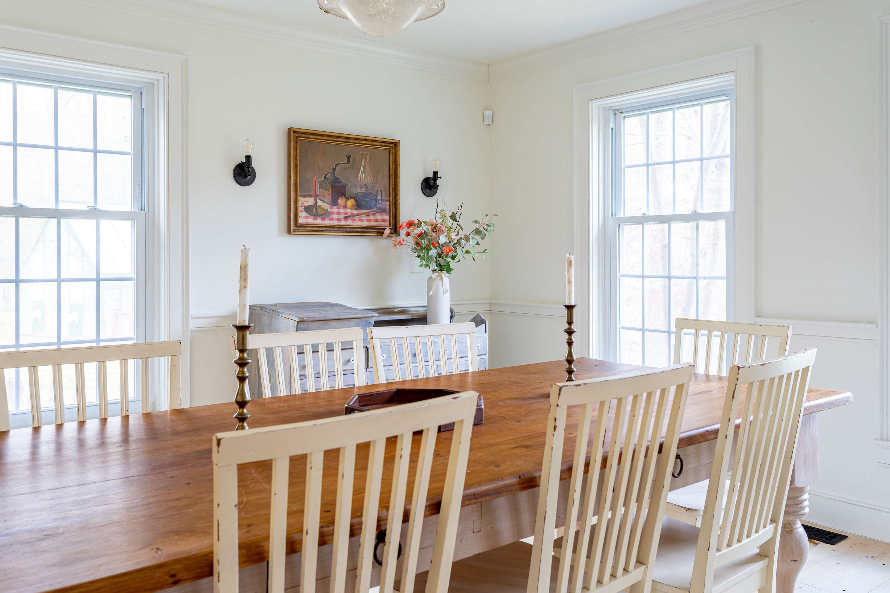 A bright dining room featuring a long wooden table with white spindle-back chairs, flanked by large windows. A vintage still-life painting hangs above a distressed blue sideboard, accented by wall sconces.