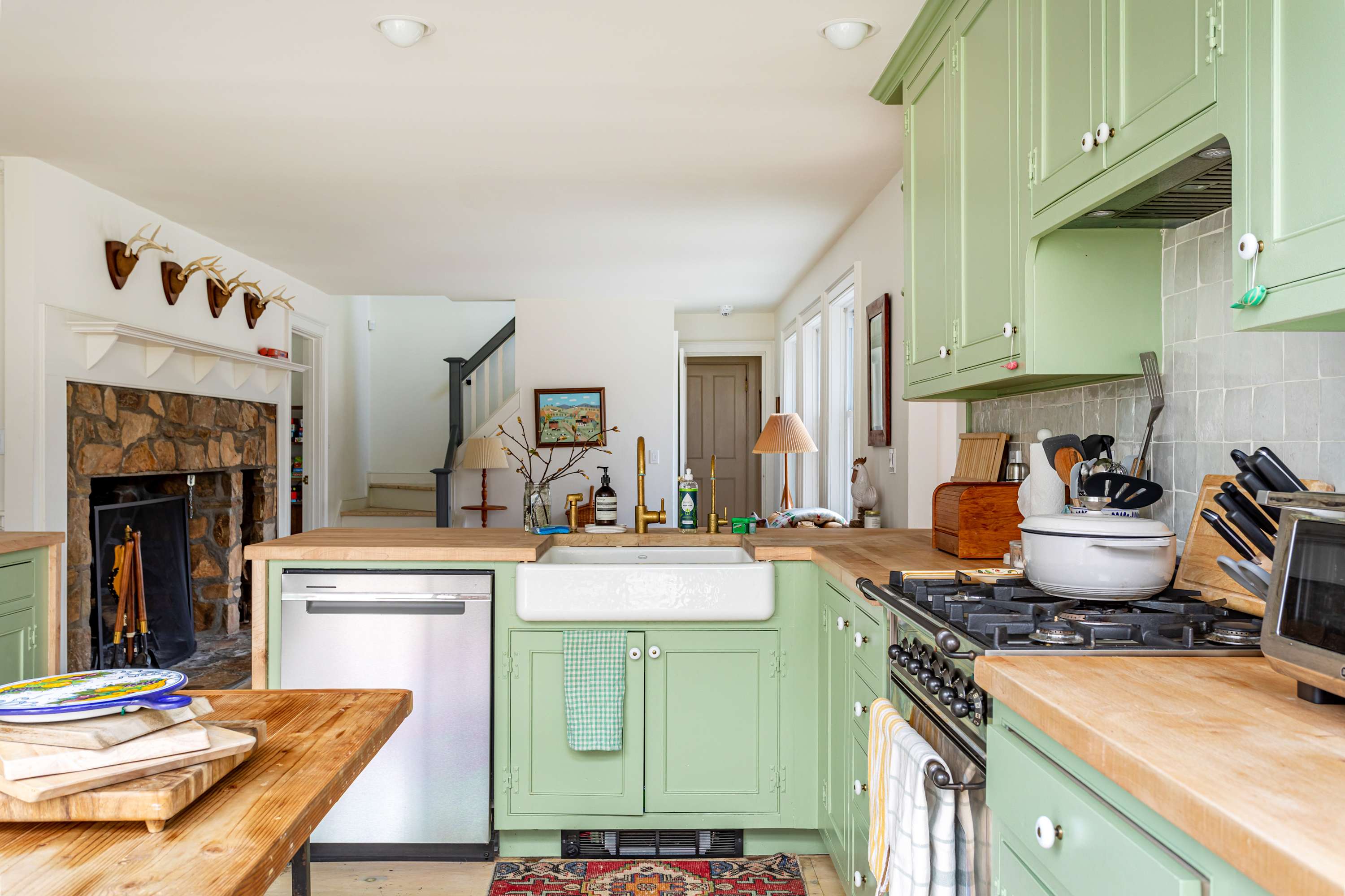 A charming kitchen with light green cabinets and butcher block countertops, featuring a white farmhouse sink with a gold faucet and a stainless steel dishwasher. A colorful rug accents the floor, and the stone fireplace of the living room is visible in the background.