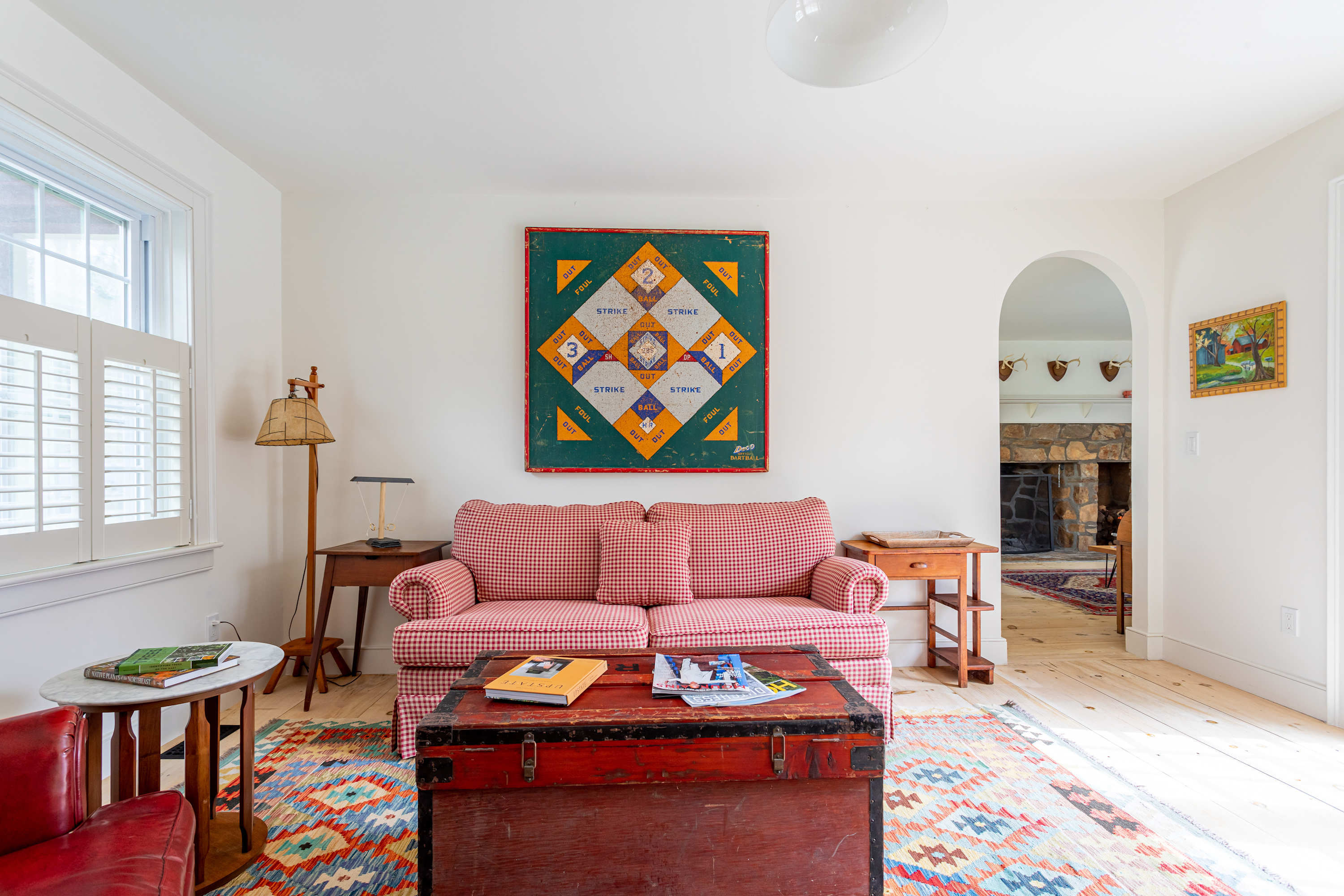 A bright sitting area featuring a red gingham sofa, a rustic red wooden trunk serving as a coffee table, and a vibrant patterned rug. An arched doorway leads to another room with a stone fireplace.
