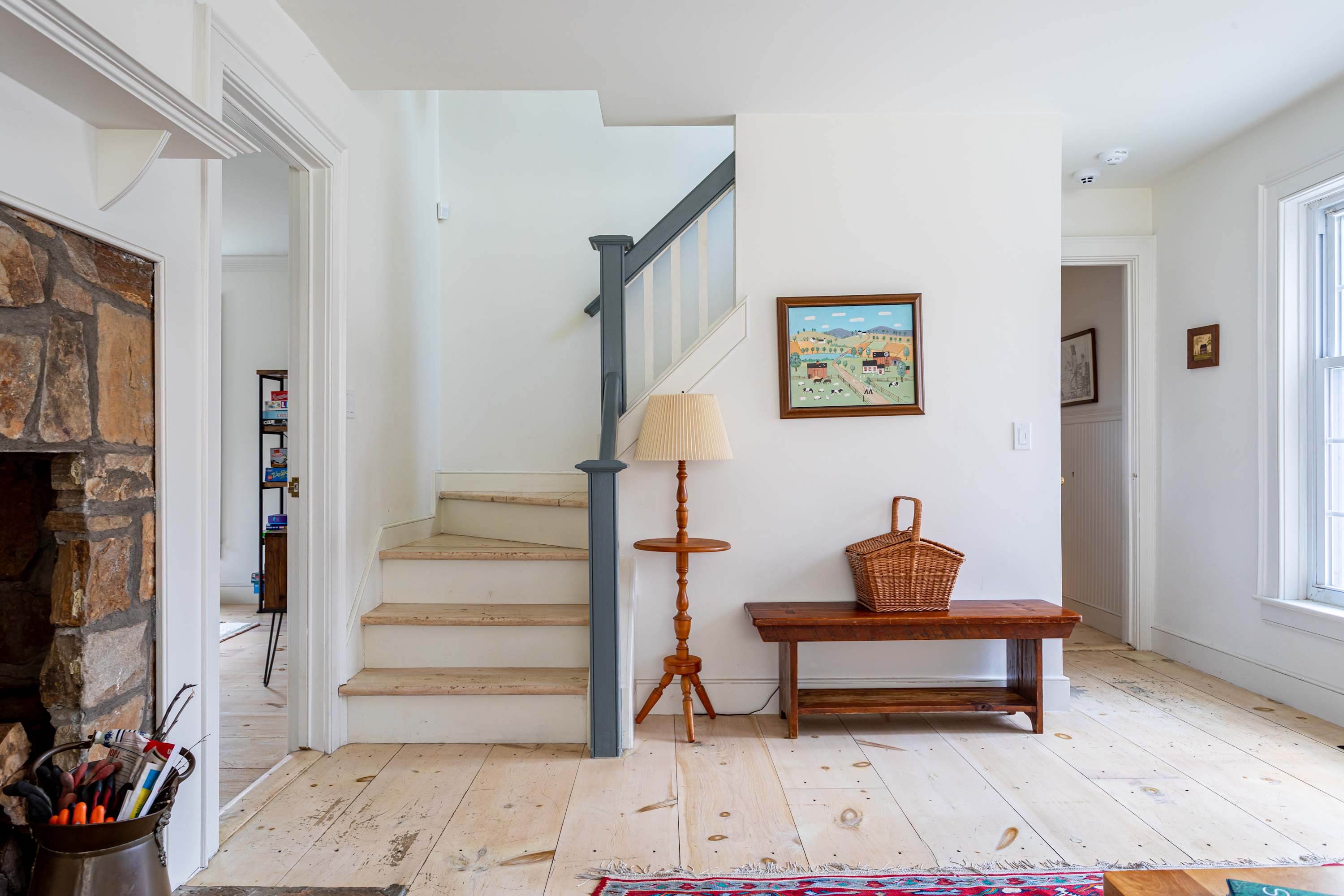 A welcoming foyer with light wide plank wood floors, featuring a staircase with a gray banister and a wooden bench with a woven basket. Artwork hangs on the wall, and a stone fireplace is partially visible to the left.