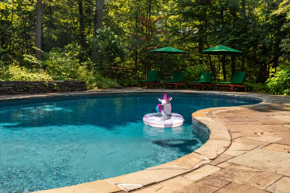 A sparkling blue saltwater pool, featuring a fun unicorn float, is elegantly curved and surrounded by a natural stone patio. The area is secluded by a dense backdrop of mature green trees, with lounge chairs and umbrellas visible in the distance.