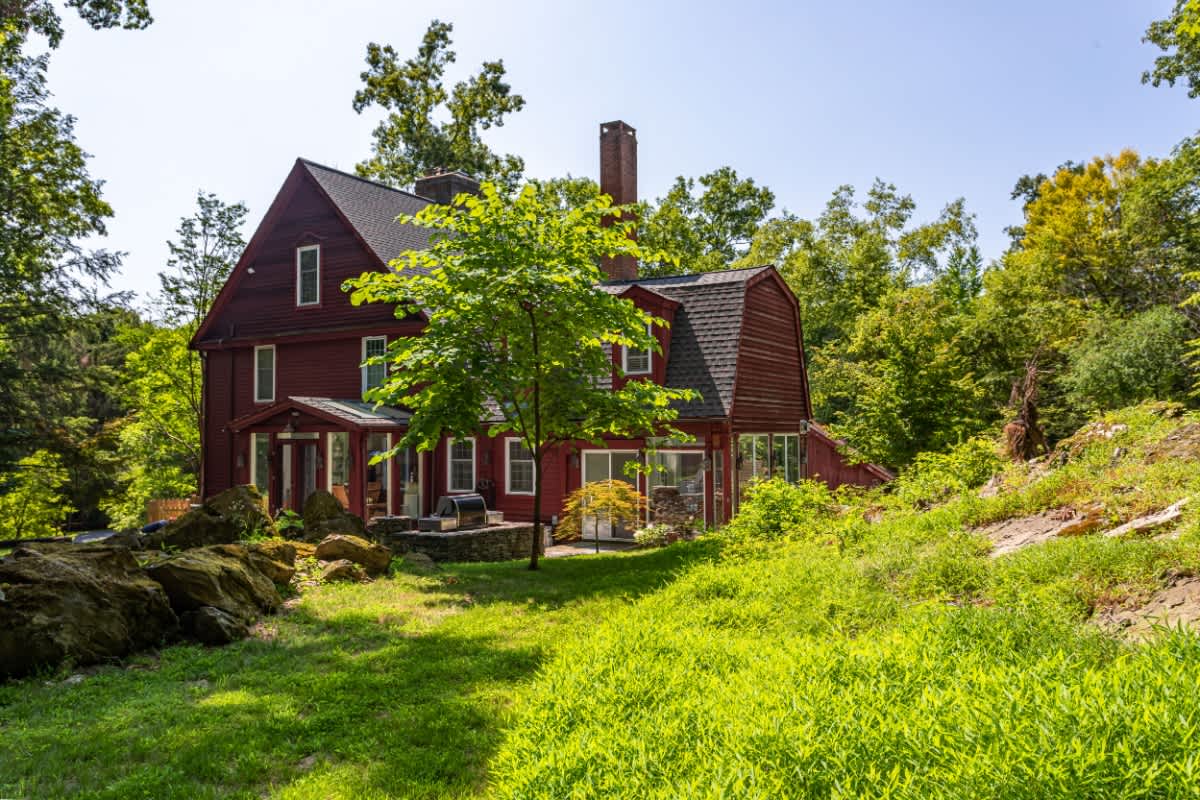 Expansive view of the red-sided house with a prominent brick chimney, nestled within a vast green lawn and surrounded by dense, mature trees under a bright sky.