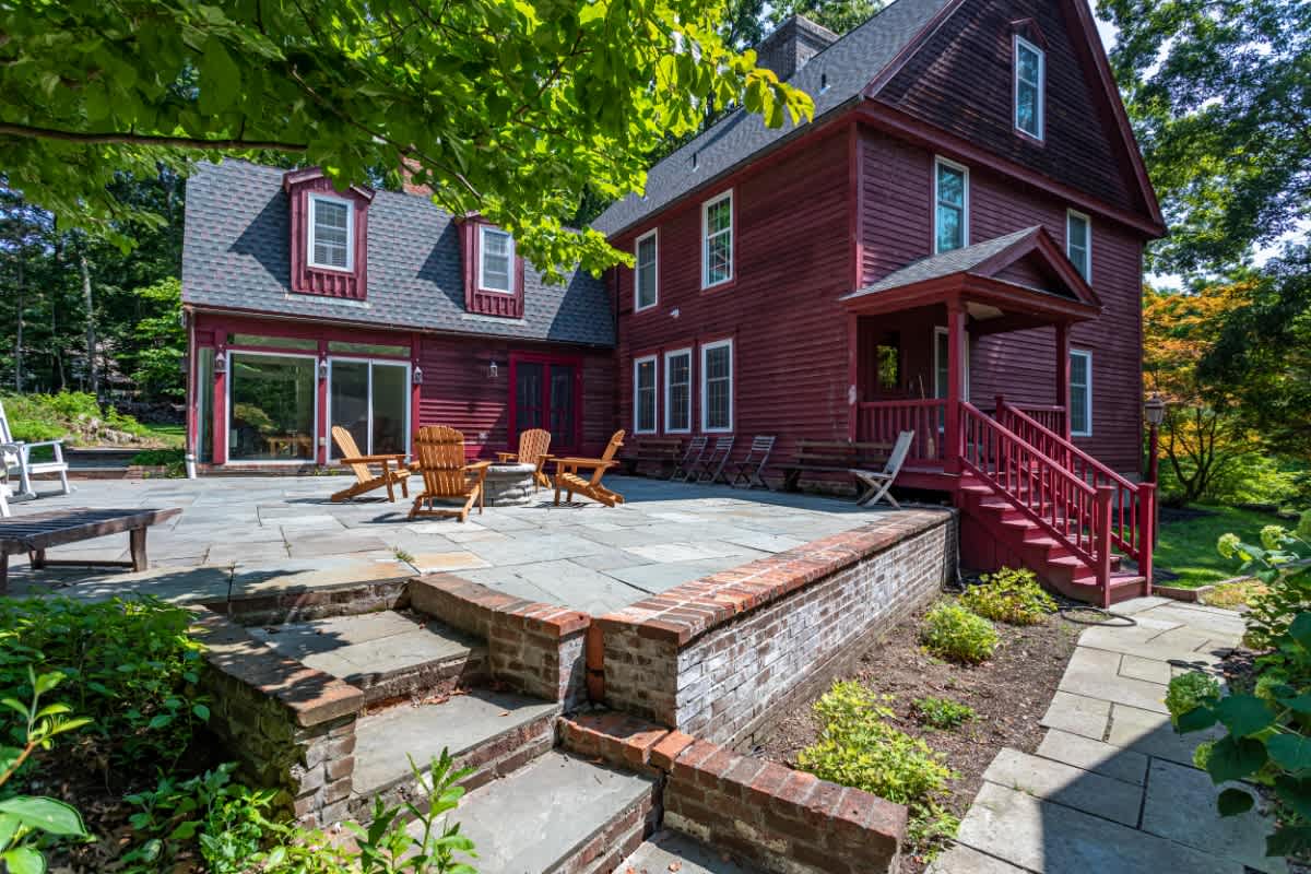 Expansive stone patio at the back of a red-sided house, featuring a central fire pit surrounded by wooden Adirondack chairs and lush green trees, perfect for outdoor gatherings.