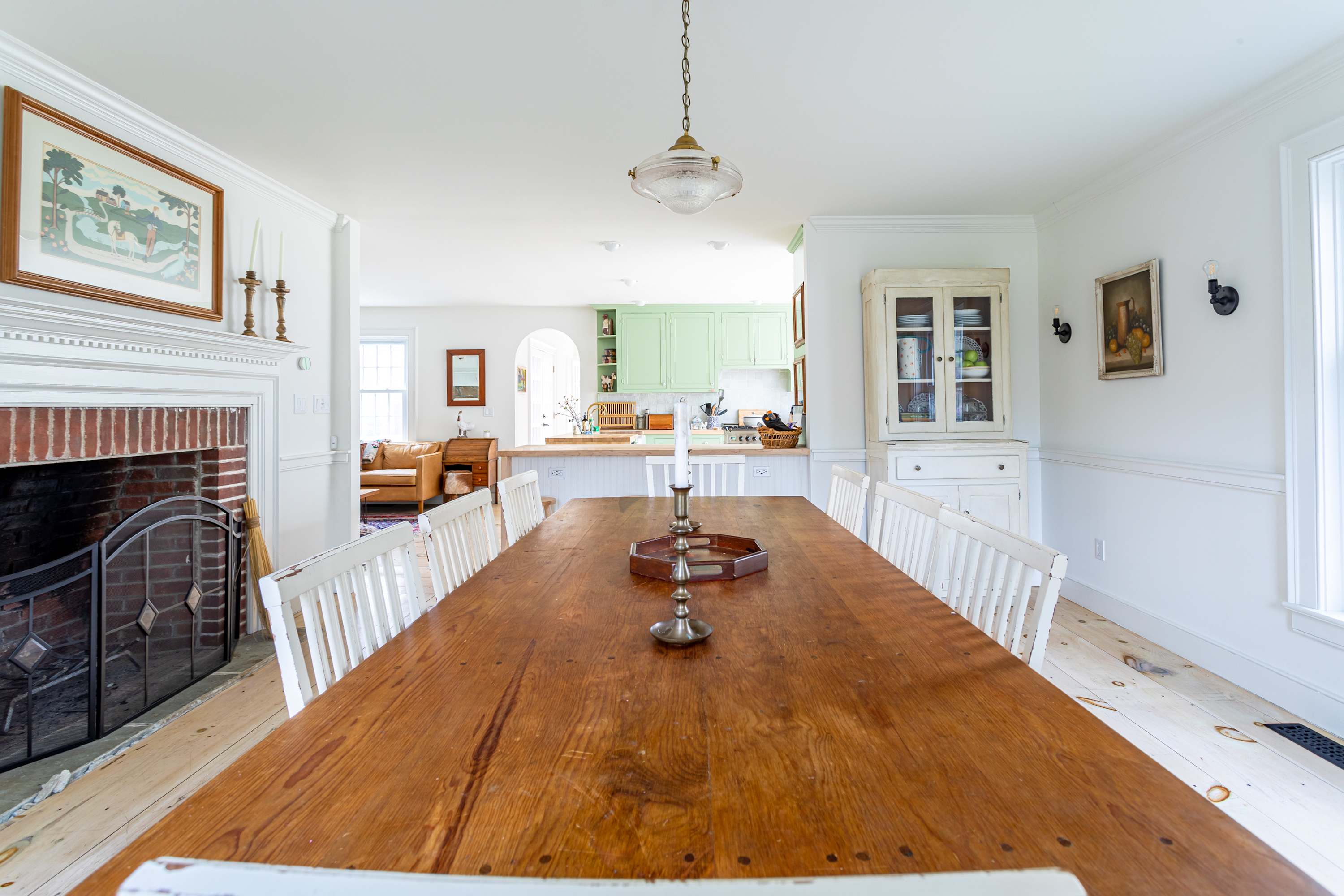 A formal dining room featuring a long, rustic wooden dining table with white chairs, illuminated by a vintage-style pendant light. A brick fireplace with a black screen is on the left, and a white hutch displays dishware on the right.