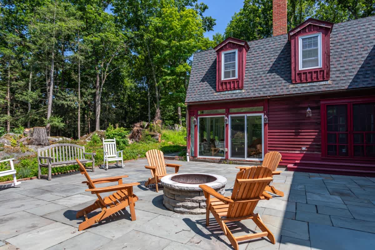 A spacious stone patio behind the red house features a circular stone fire pit surrounded by comfortable wooden Adirondack chairs, ideal for outdoor relaxation. The area is bordered by lush trees and offers a view of the house's red siding and dormer windows.