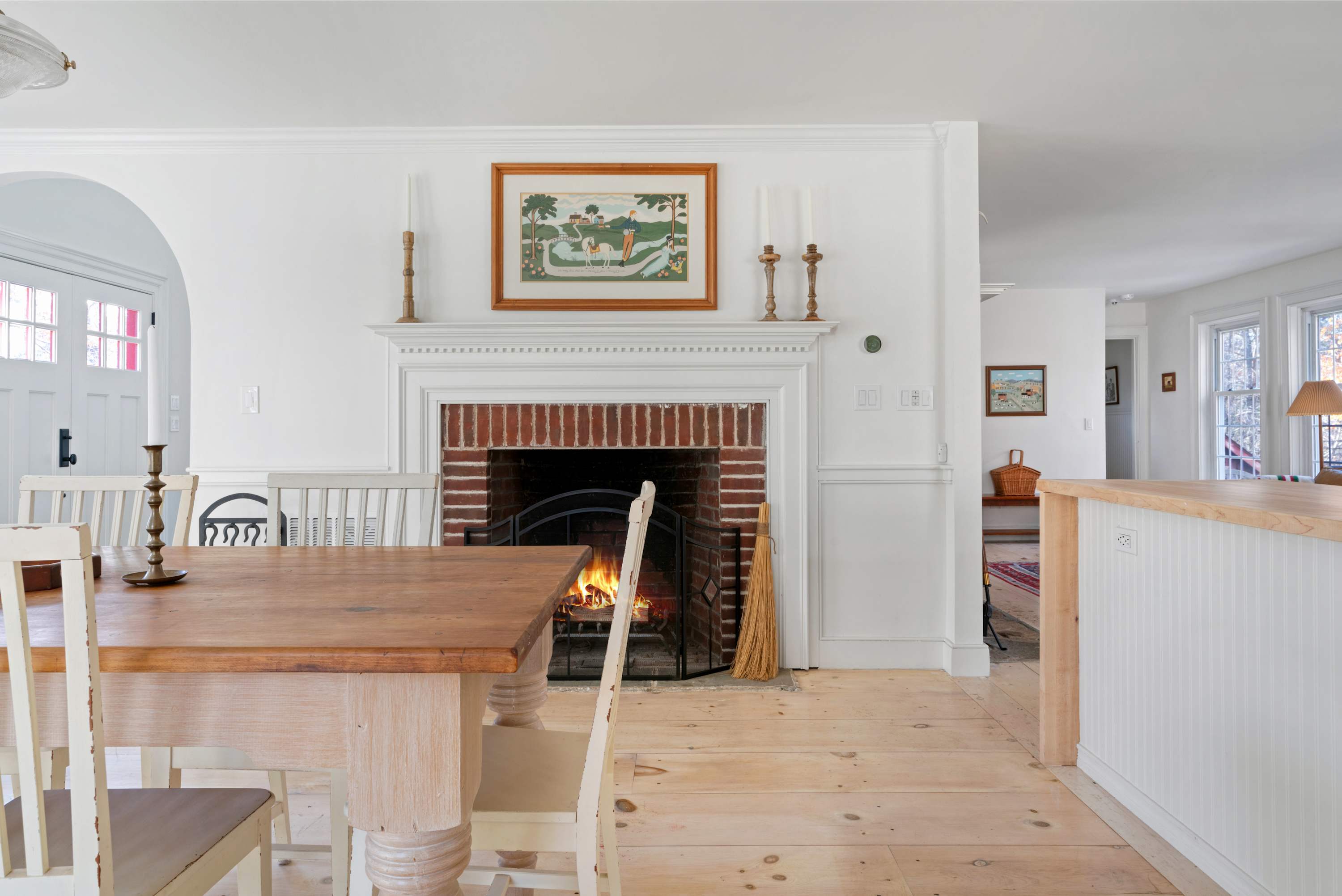 A charming dining area features a light wood dining table with white chairs, positioned in front of a brick fireplace with a white mantel and a burning fire. Light wood floors and white walls create a bright and inviting atmosphere, with an open archway leading to another room.