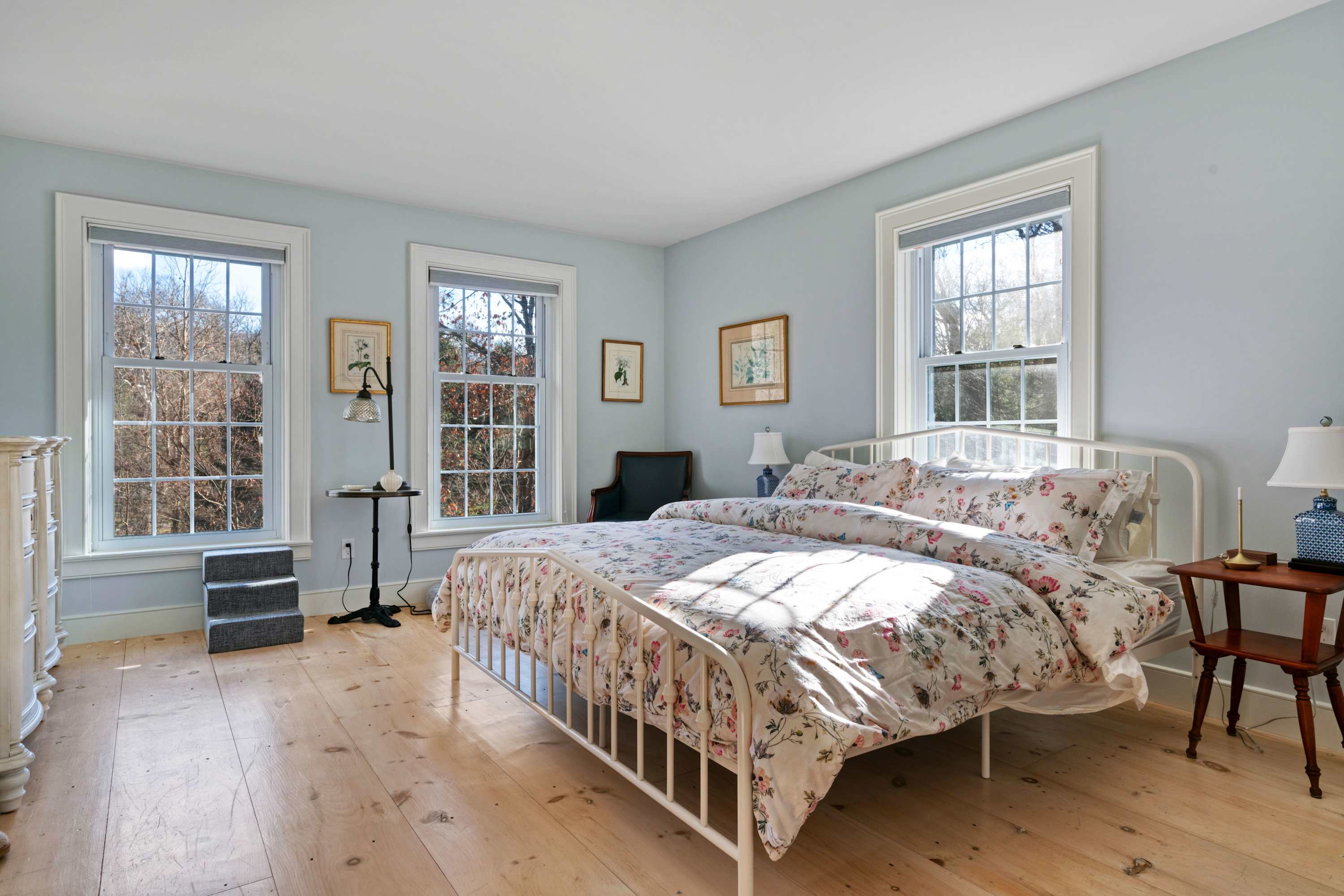 A serene bedroom with light blue walls and wide plank wood floors, featuring a white metal bed frame dressed with a floral duvet. Multiple windows provide ample natural light, and a small wooden nightstand holds a lamp.