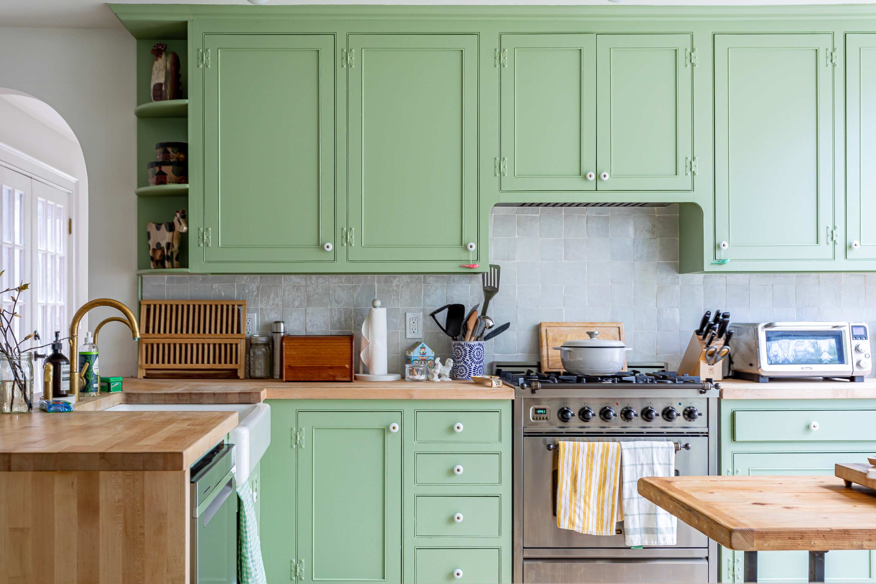 A detailed view of the kitchen counter showcasing light green shaker cabinets, a light gray tiled backsplash, and a stainless steel range with various cooking utensils. Wooden cutting boards and small appliances are neatly arranged on the butcher block countertop.