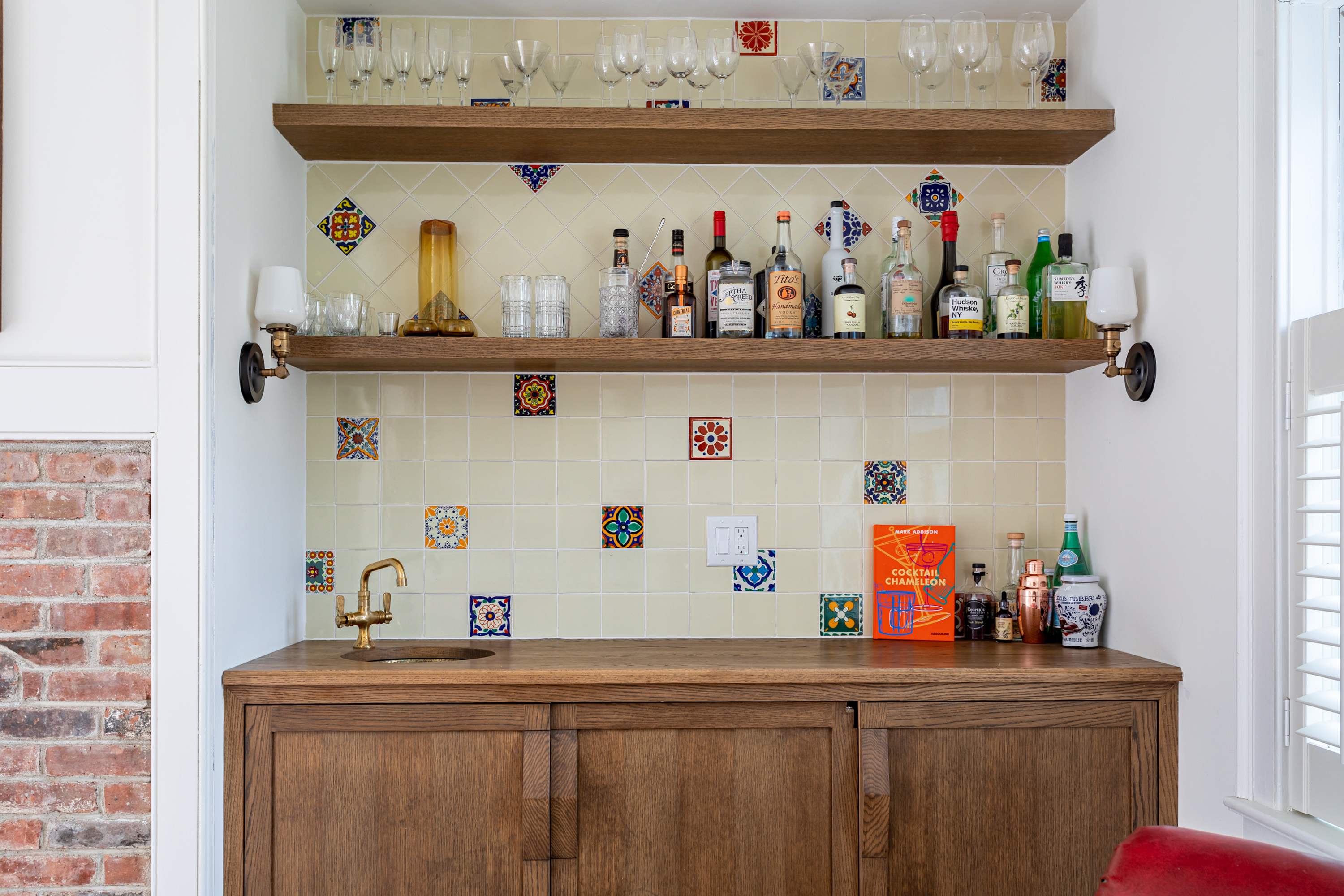 A stylish wet bar featuring a wooden base cabinet with a brass sink and faucet, complemented by a colorful tiled backsplash. Open wooden shelves display various bottles and glassware.