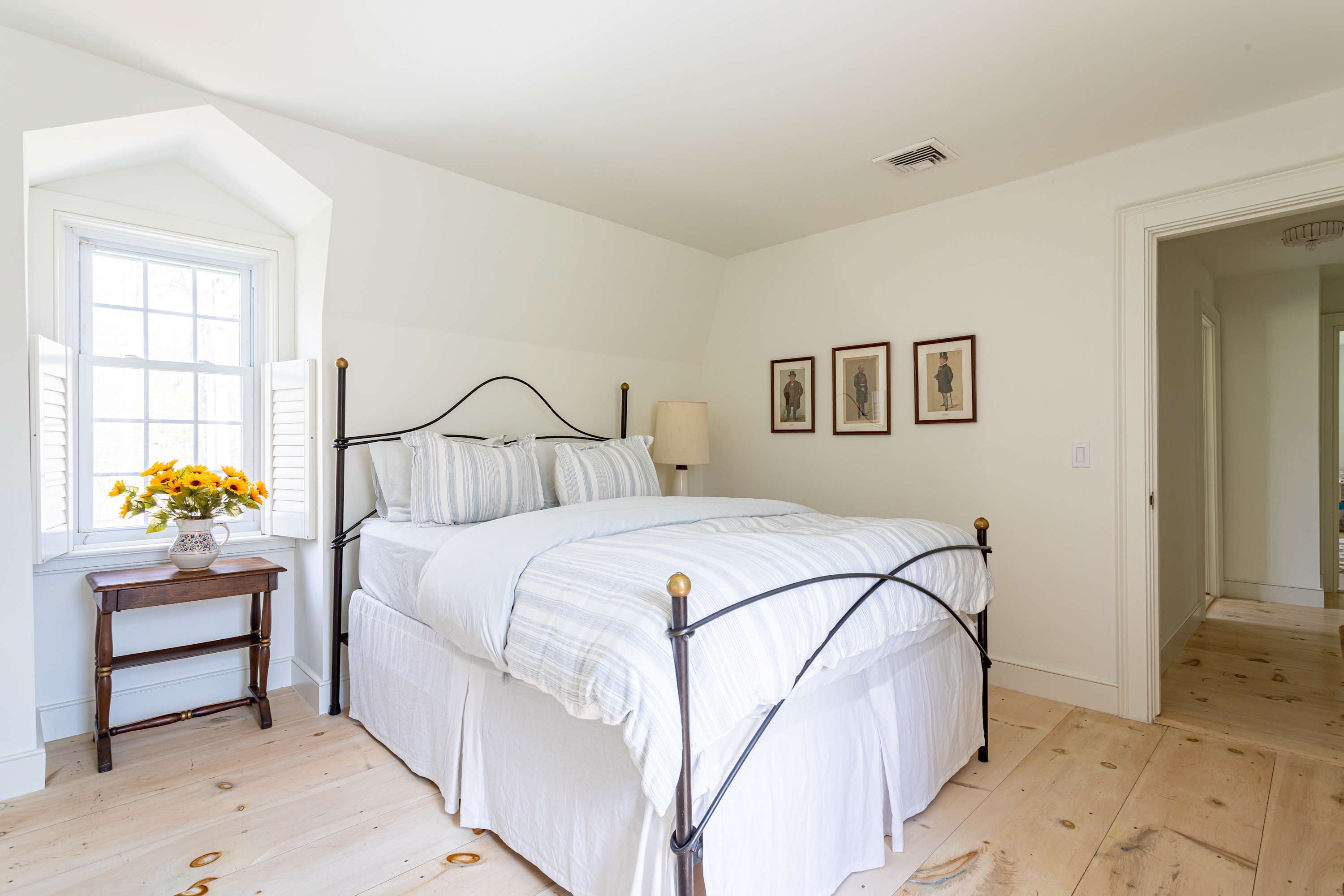 Bright bedroom with white walls and light wood floors, featuring a black and brass metal bed frame with striped white bedding. A small wooden side table with a vase of yellow flowers sits next to a window with white shutters.