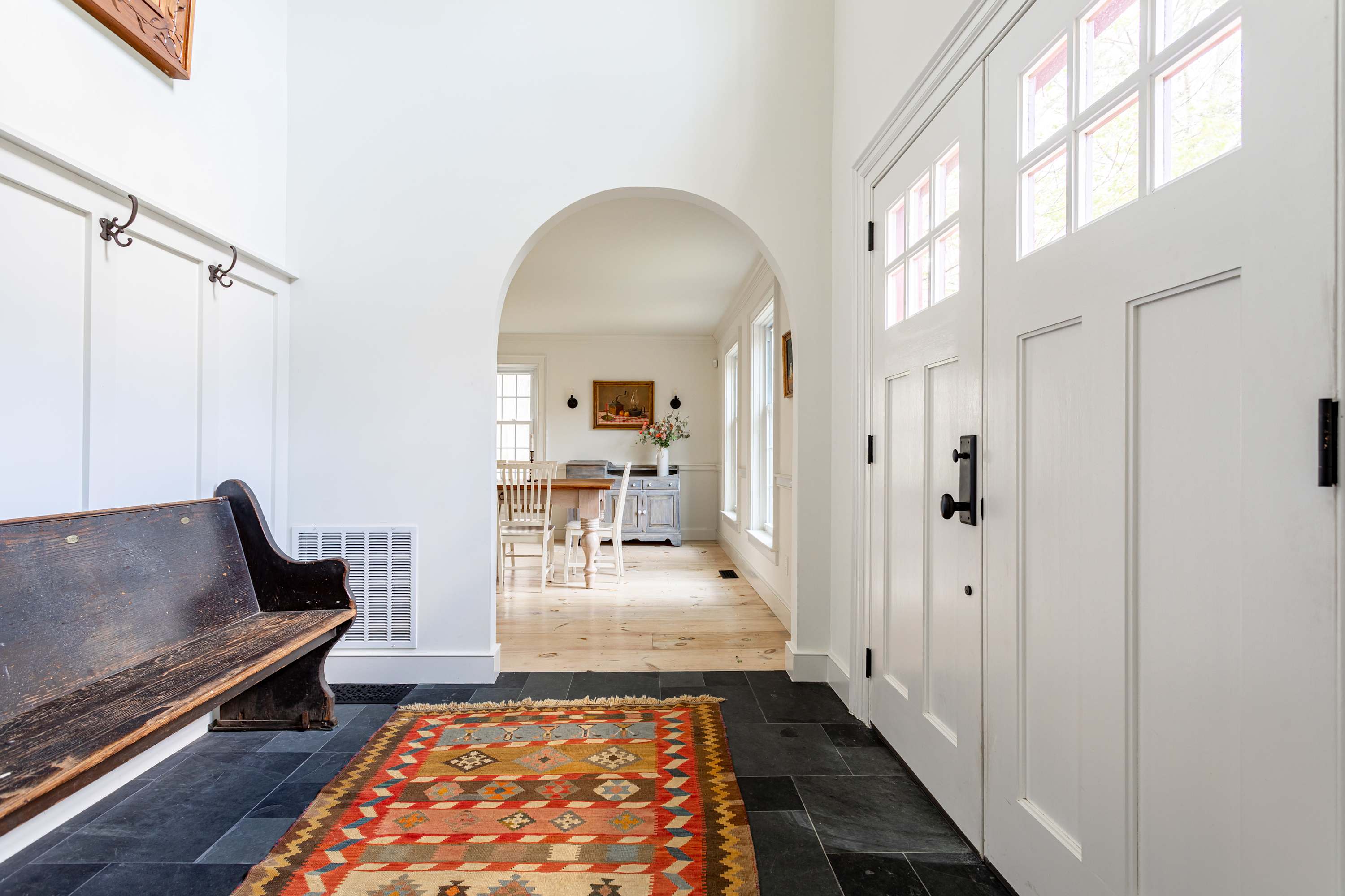 A welcoming foyer with dark slate tile floors and a colorful geometric rug, featuring a rustic wooden bench and white wainscoting. A white front door with glass panes is on the right, and an arched doorway leads to a dining area.