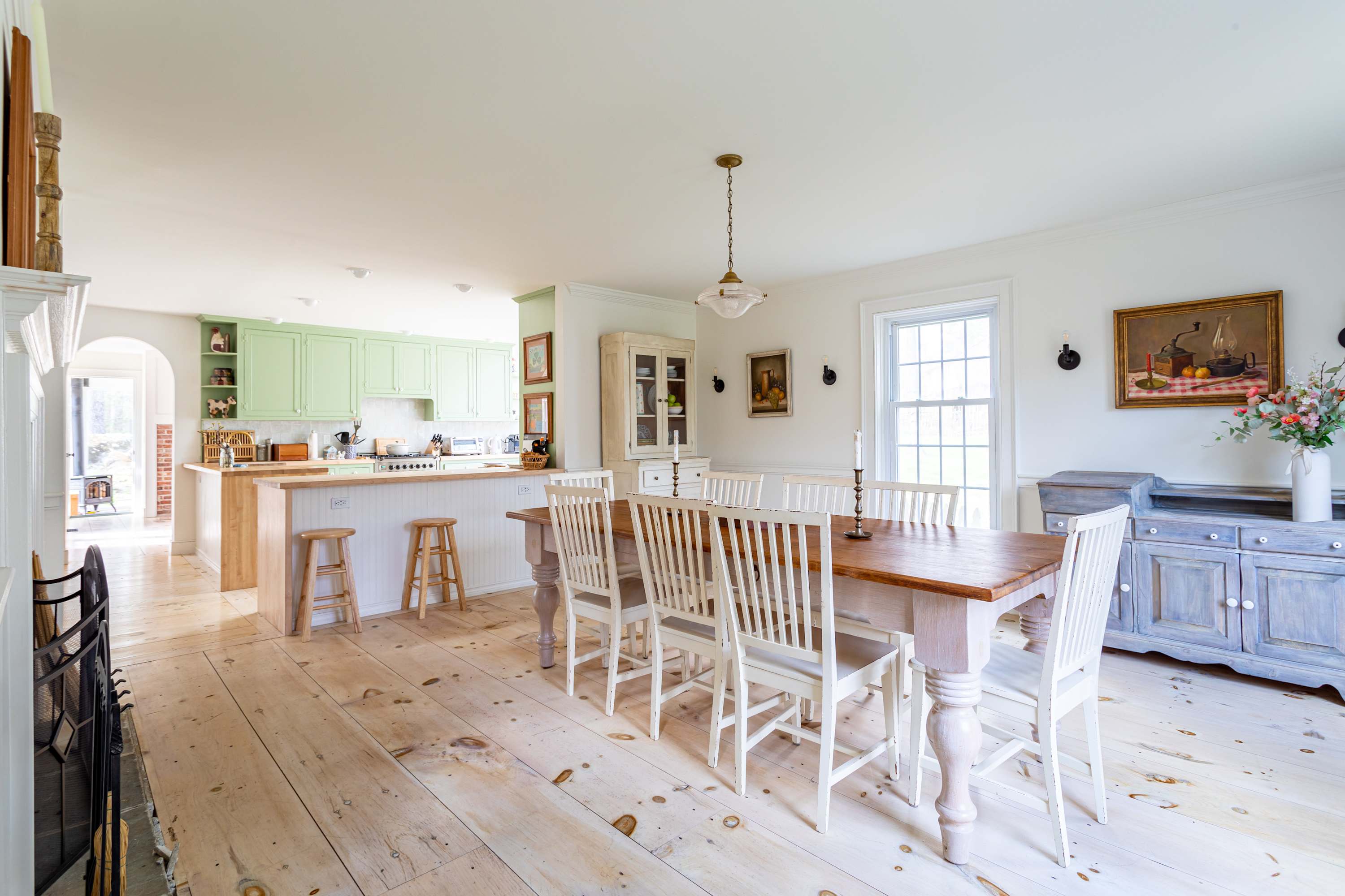 An open-concept dining area with a long wooden table and white chairs, leading into a kitchen with light green cabinetry and a wooden island with stools. Light-colored wide plank wood floors extend throughout the space.