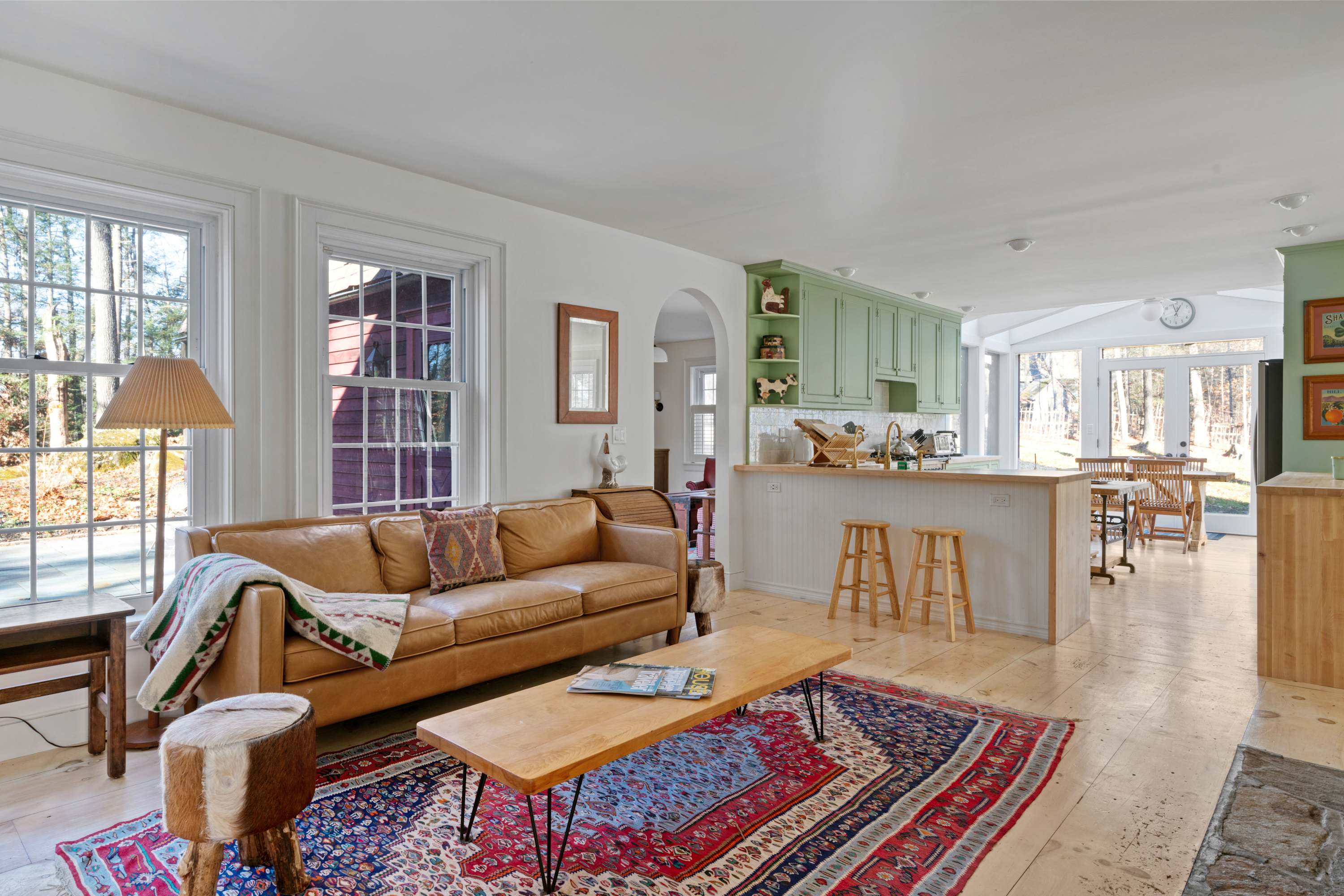 An expansive open-concept living space with a tan leather sofa, wooden coffee table, and a colorful rug, leading into a kitchen area with light green cabinets and a breakfast bar. Large windows offer views of the surrounding trees.