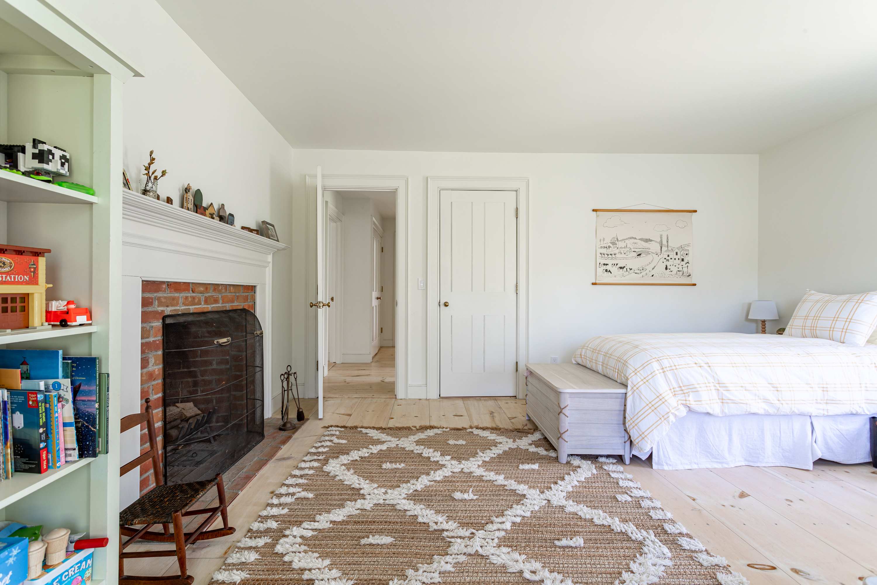 Spacious white-walled bedroom with light wood floors, showing one of the twin beds, a brick fireplace with a white mantel, and a built-in bookshelf. A patterned rug and a rocking chair are also visible.