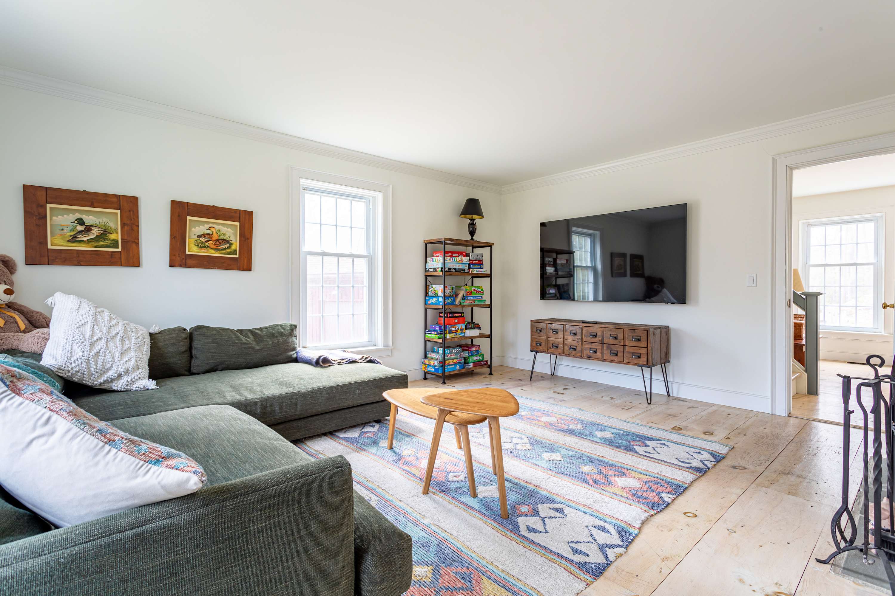 A cozy living room with a large green sectional sofa, a colorful patterned rug, and a wall-mounted television above a rustic wooden console. A tall bookshelf filled with games stands next to a window.