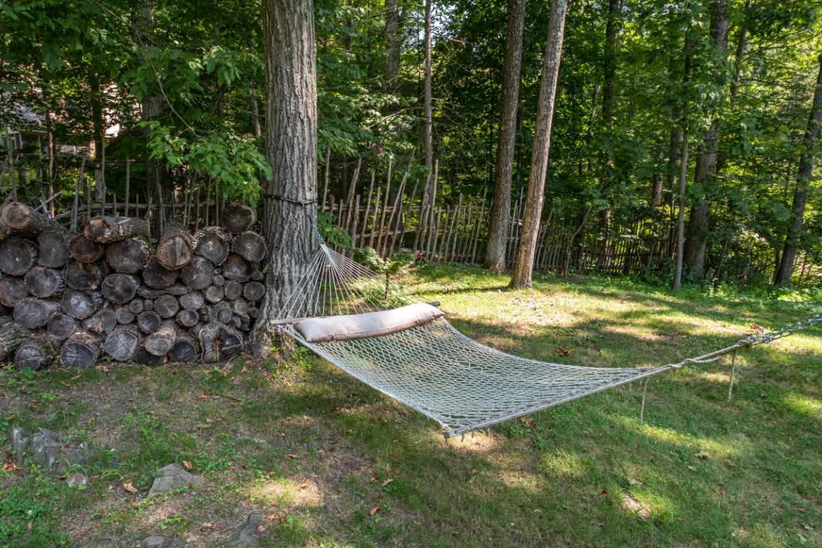 A comfortable rope hammock is strung between two mature trees in a shaded, grassy yard, with a neatly stacked pile of firewood nearby and a rustic fence in the background.