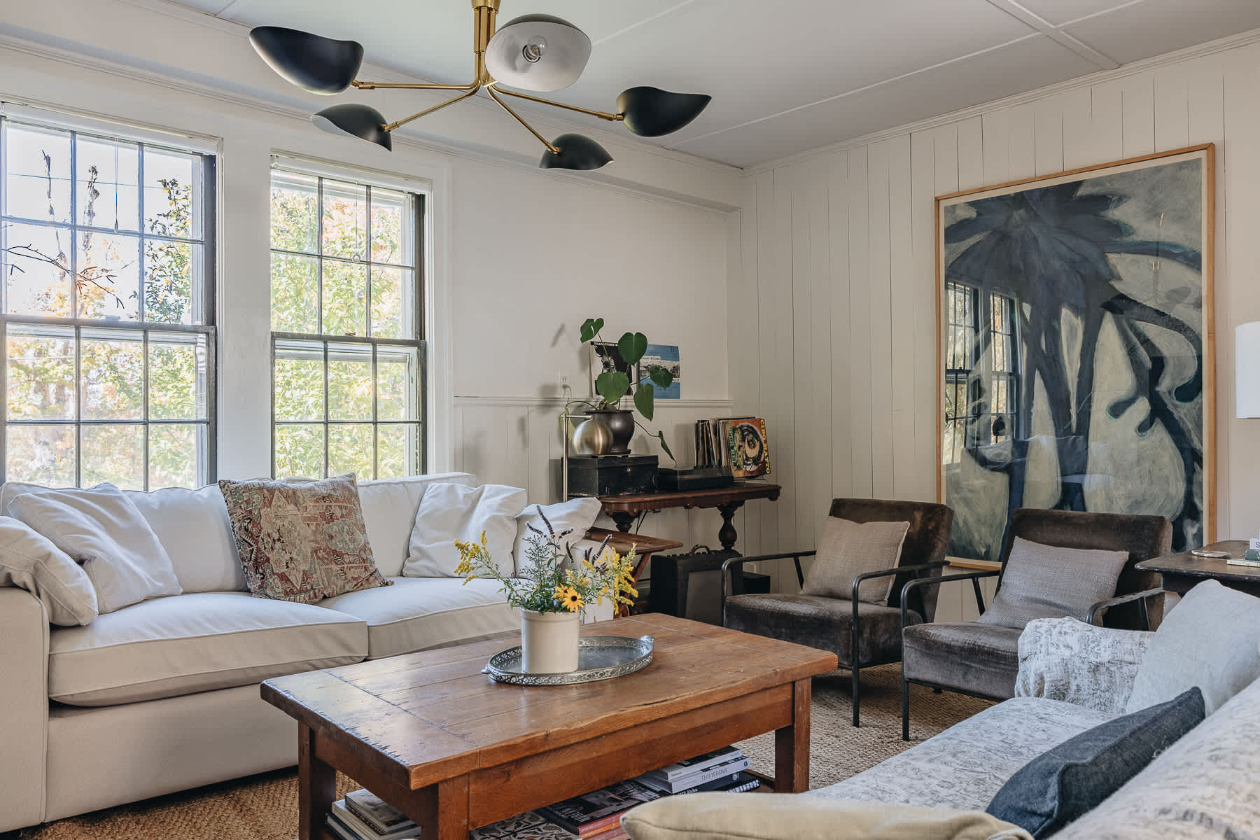 Bright living room with white paneled walls, a large white sofa, two armchairs, and a rustic wooden coffee table, illuminated by a modern chandelier.