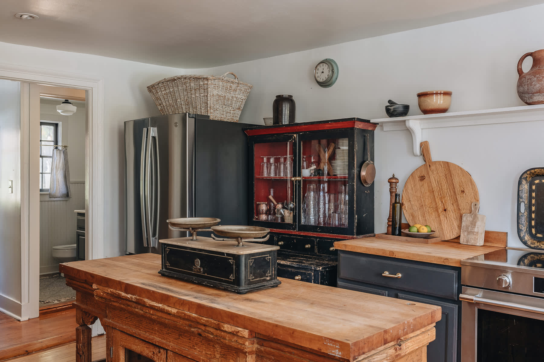 Kitchen view showcasing a stainless steel refrigerator, a large rustic wooden island with a vintage scale, and a distressed black and red display cabinet.