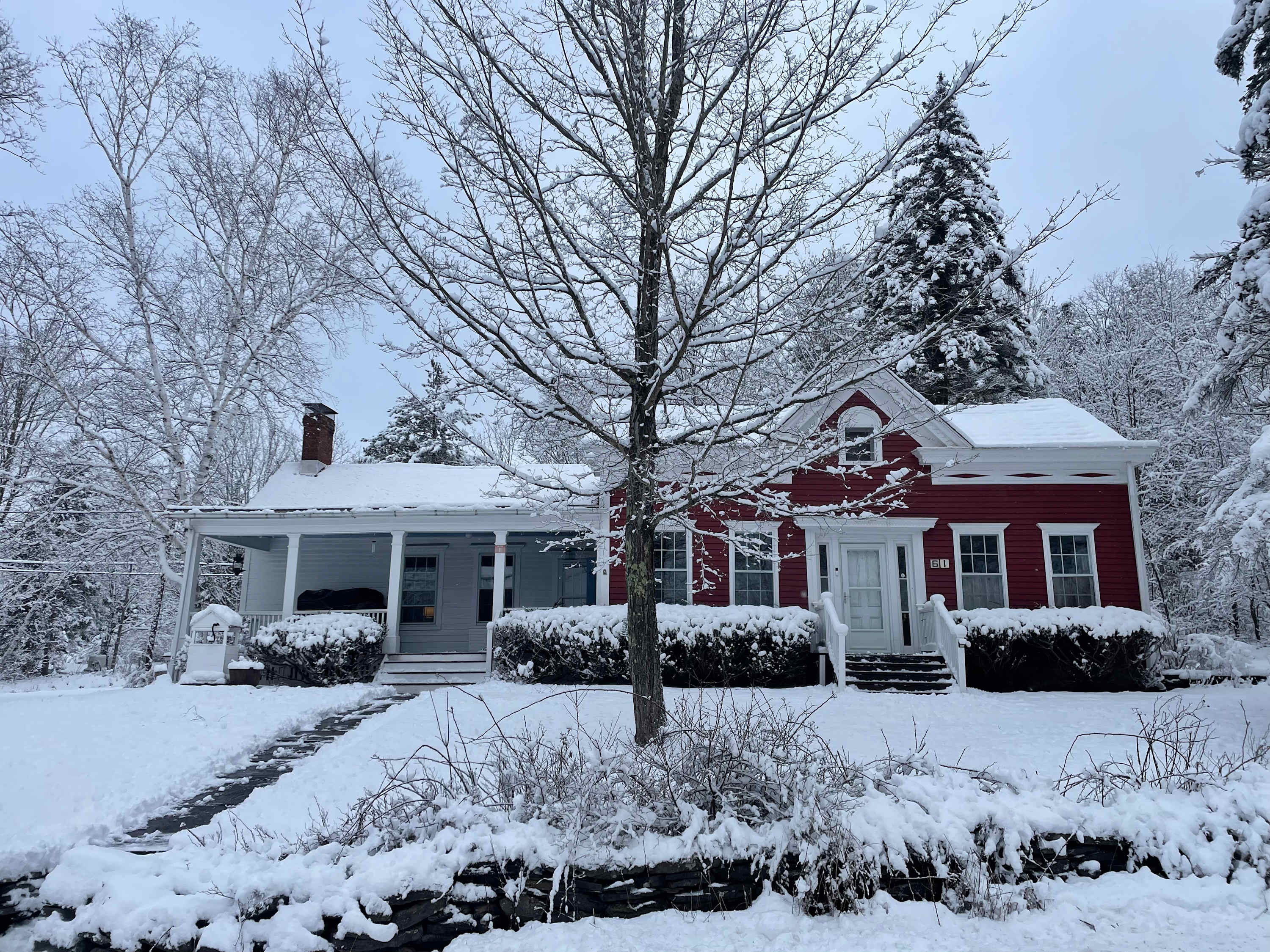 A charming red farmhouse with white trim and a covered porch is blanketed in fresh snow, with a snow-dusted path leading to the front door. Bare trees and evergreen bushes are also covered in snow, creating a serene winter scene.