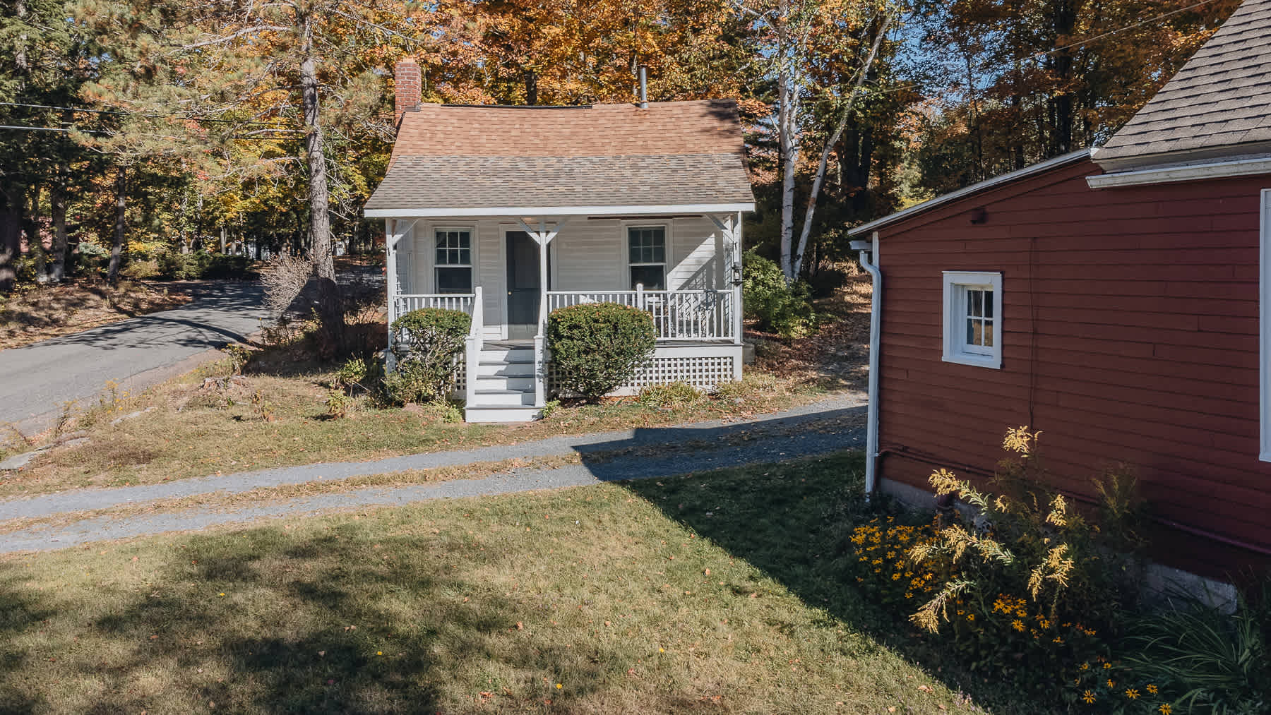 An aerial view showcases a charming white cottage with a covered front porch and a red-sided outbuilding, surrounded by vibrant autumn trees. A gravel driveway leads up to the house, with green grass and yellow flowers in the foreground.