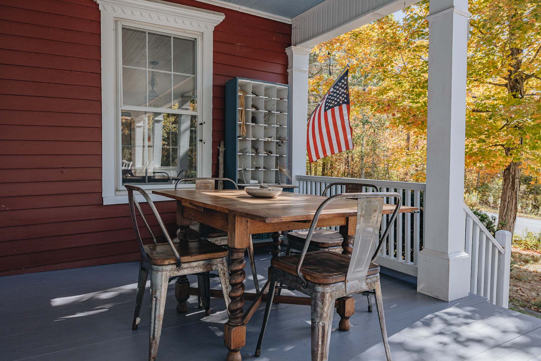 A covered porch with a wooden dining table and metal chairs, set against the red exterior of the house. An American flag hangs nearby, and a blue cubby shelf is visible through a window.