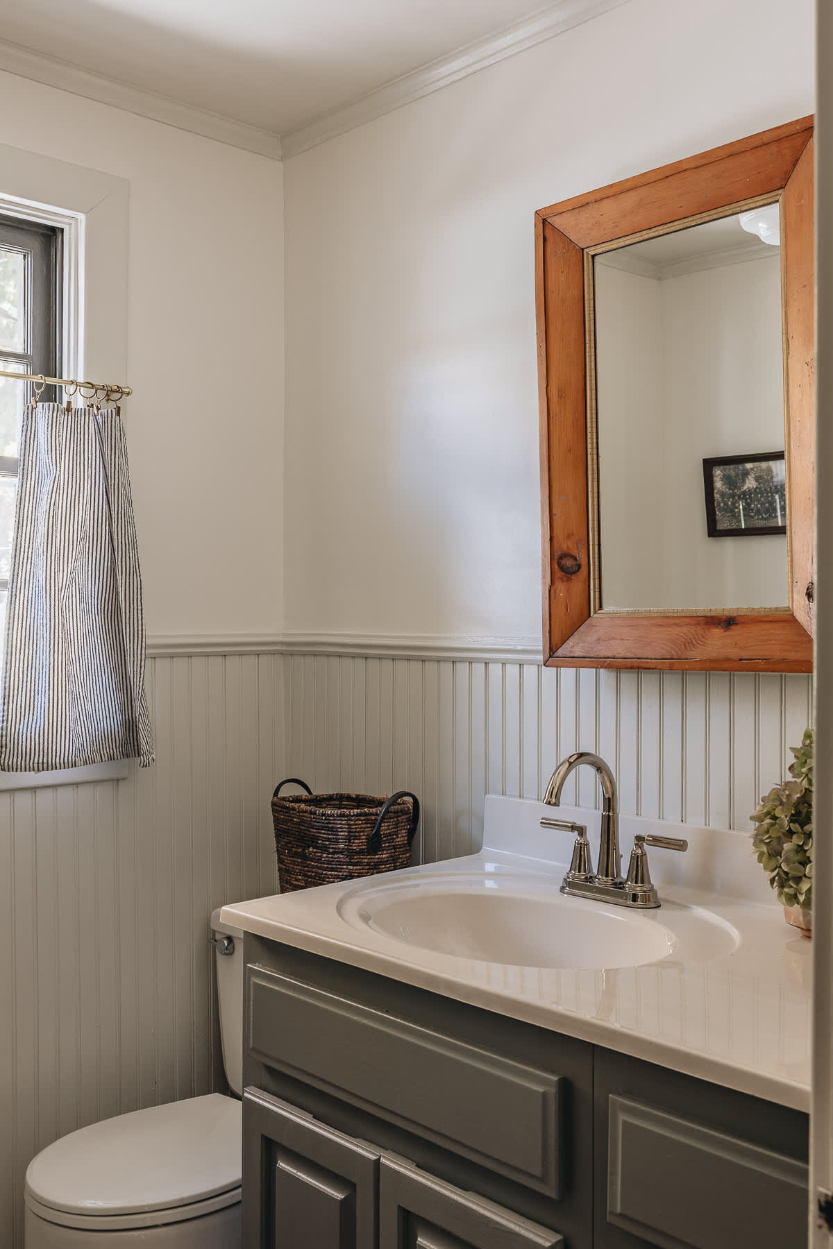A cozy bathroom features white beadboard wainscoting and light walls, with a dark gray vanity supporting a white sink and chrome faucet. A rustic wooden mirror hangs above, reflecting a picture, and a small window with a striped curtain is next to the toilet.