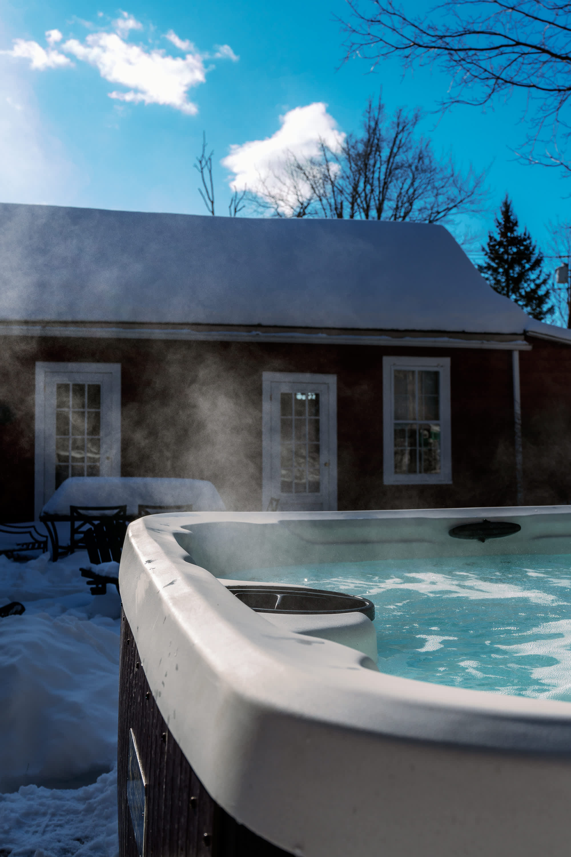 A steaming hot tub sits in a snowy backyard, with the red farmhouse visible in the background under a bright winter sky. The warm steam contrasts with the cold, snowy surroundings.