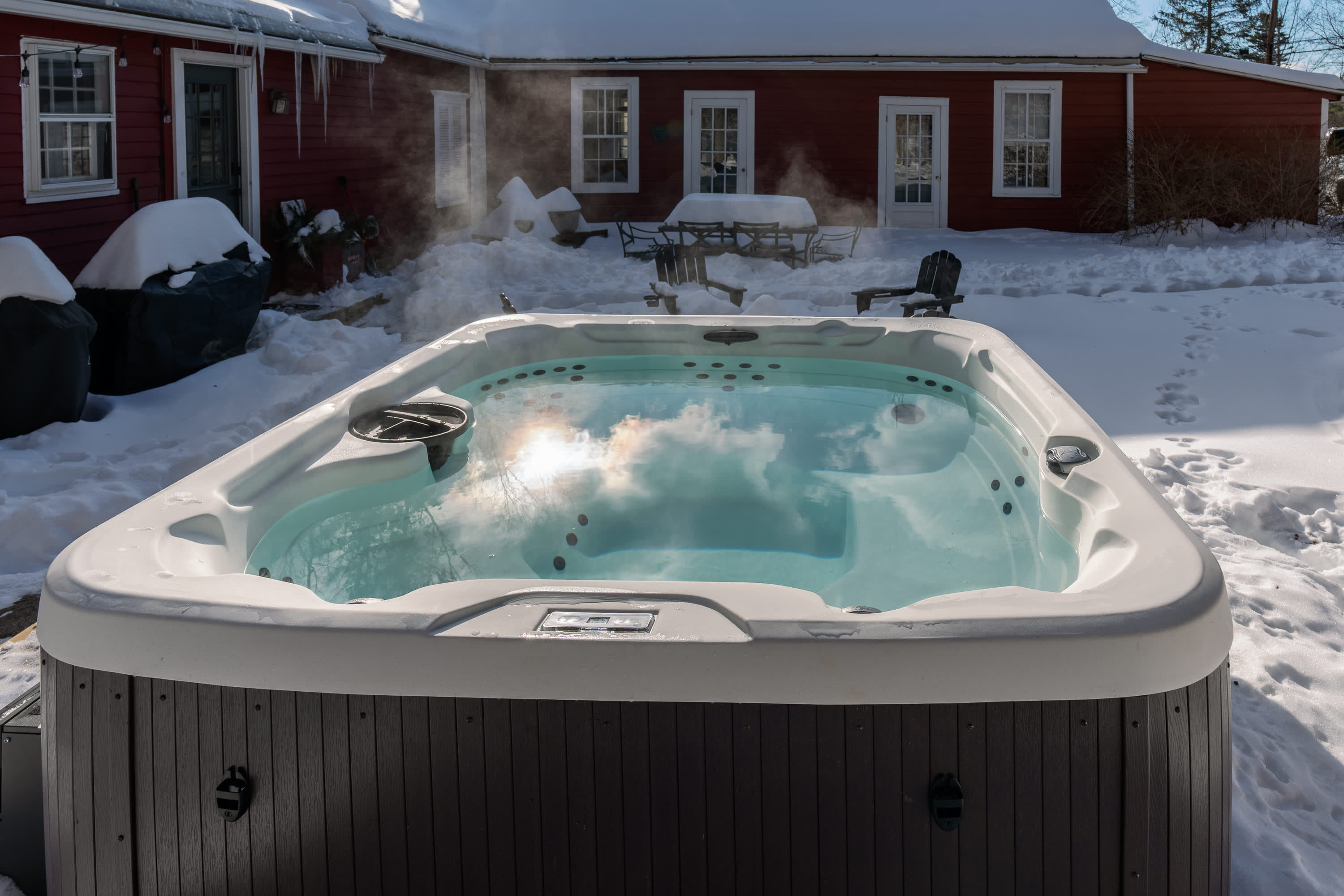 Outdoor hot tub in a snowy backyard, reflecting the sky, with a red house and outdoor furniture covered in snow in the background.