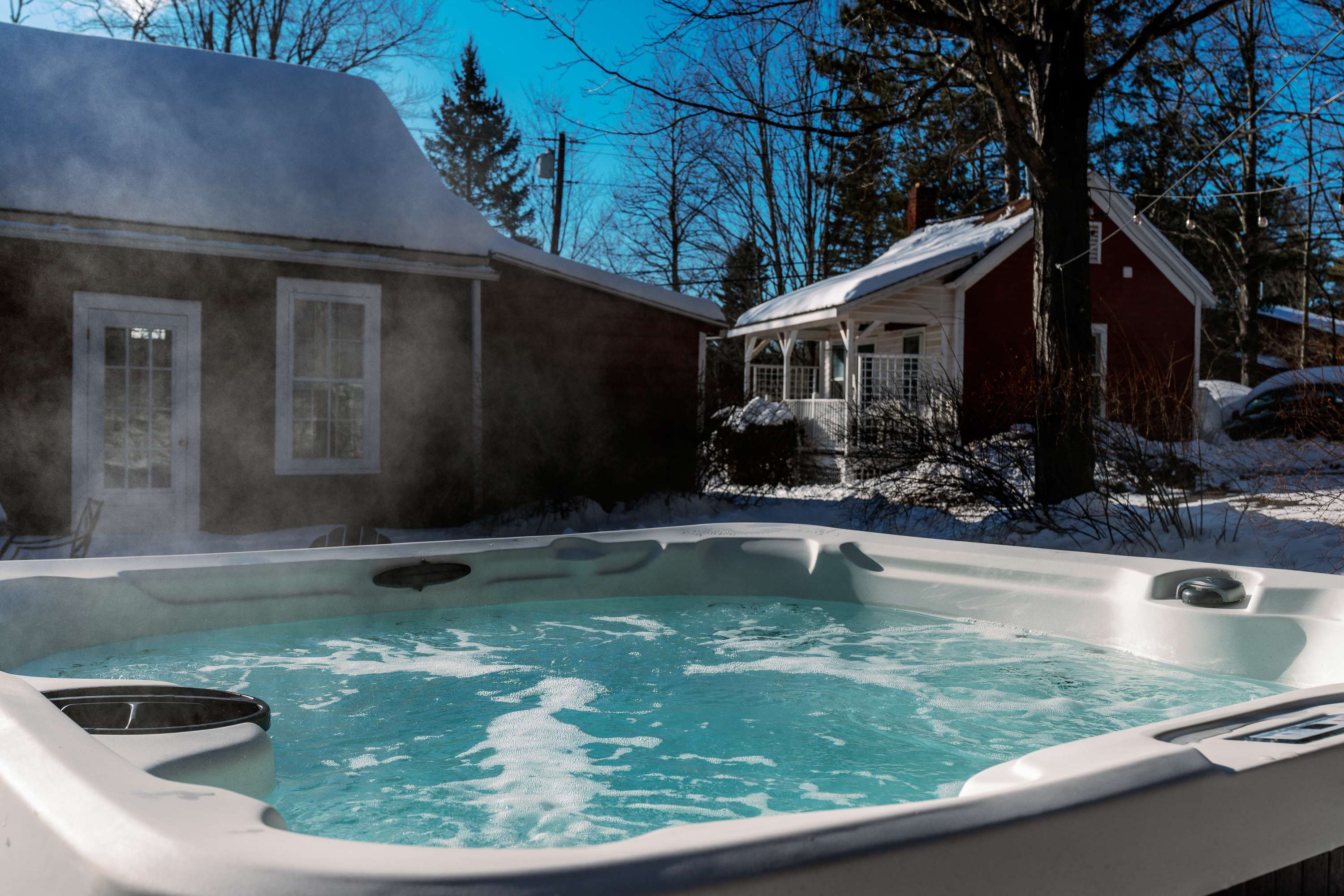 Outdoor hot tub steaming in a snowy winter landscape, with a red house visible in the background, suggesting a relaxing cold-weather retreat.