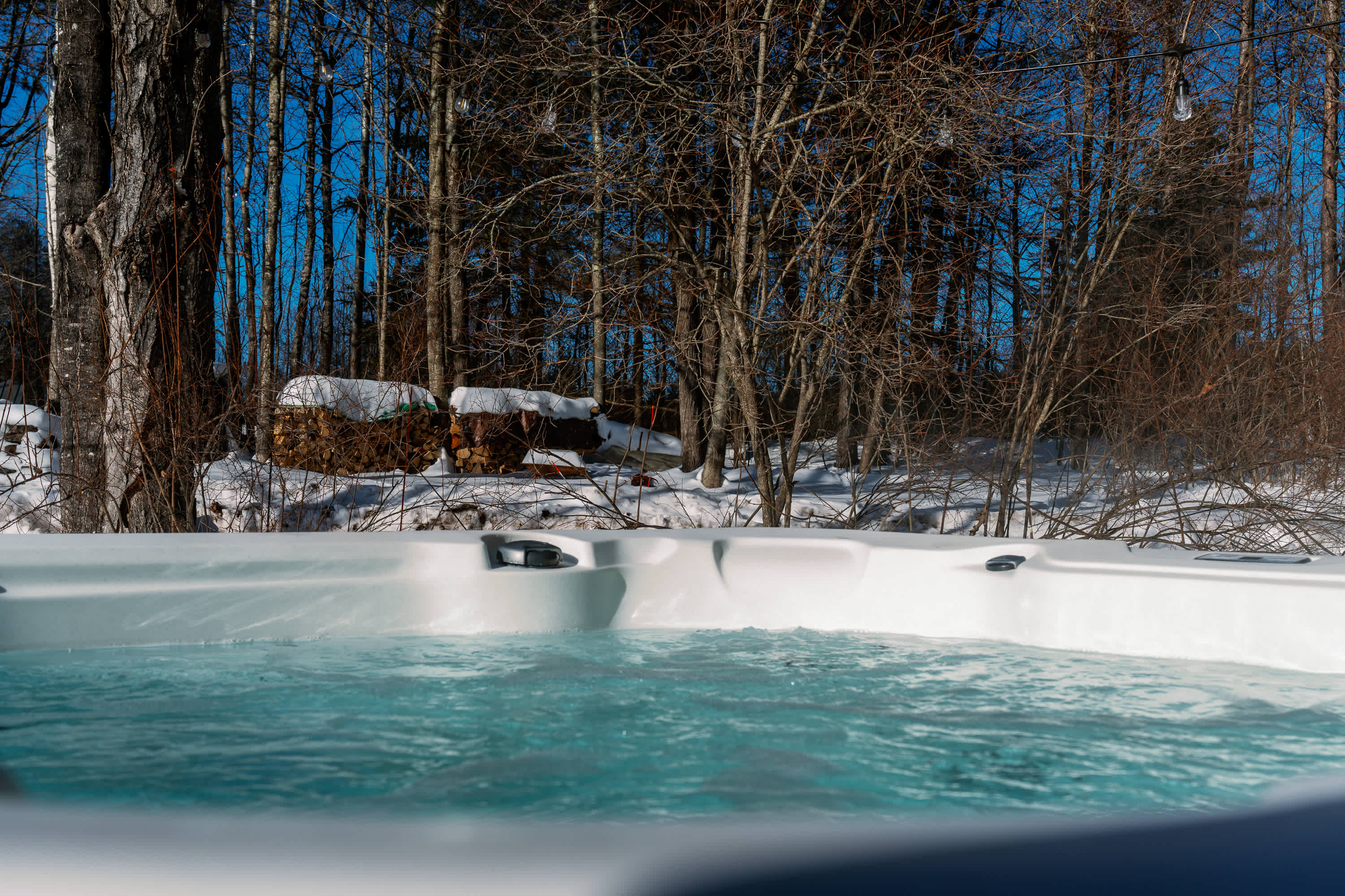 A close-up view of a steaming hot tub with clear blue water, set against a snowy winter landscape with bare trees under a bright blue sky. The scene emphasizes relaxation in a cold environment.