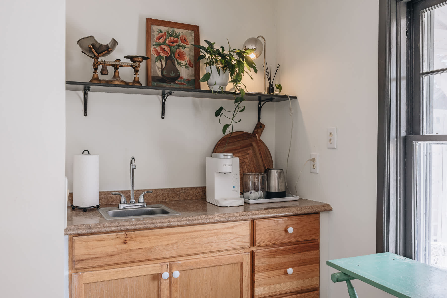 A compact kitchenette features light wood cabinetry and a white countertop with a stainless steel sink and chrome faucet. A coffee maker and kettle are neatly arranged on a tray, beneath a black floating shelf adorned with plants and decorative items.