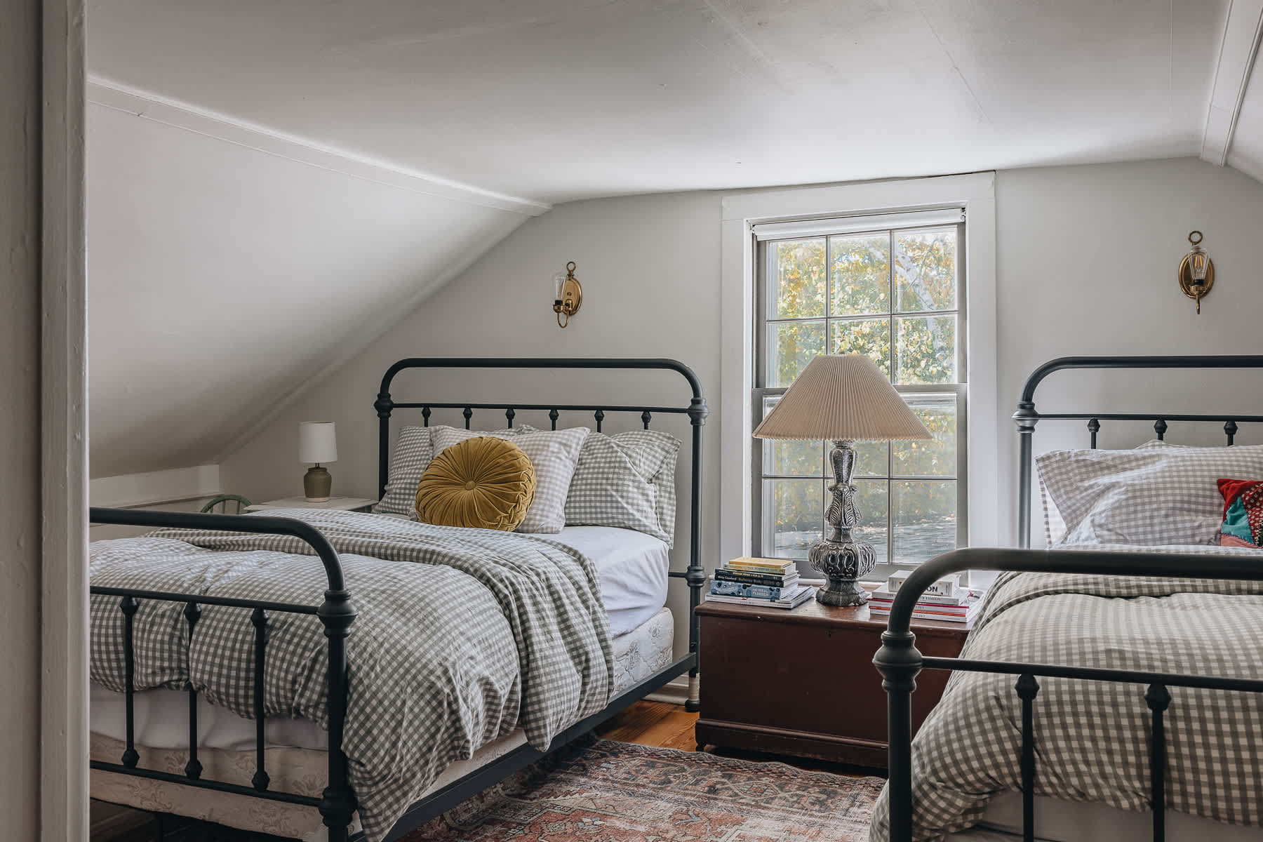 A bright bedroom with sloped ceilings and light walls features two black metal-frame twin beds dressed in gray gingham bedding. A wooden chest serves as a nightstand between them, holding a large lamp and books, with a window offering natural light.