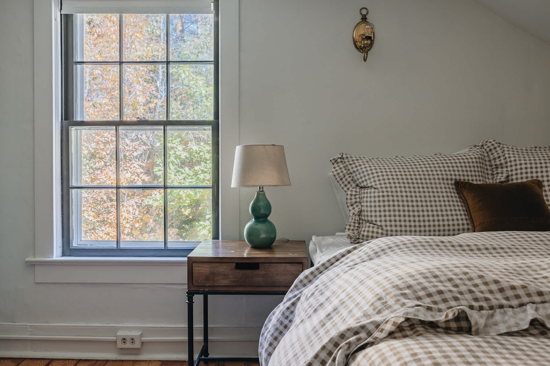 A cozy bedroom features a bed with light brown gingham bedding and a dark wood nightstand with a green ceramic lamp. A large window with black trim offers a view of autumn foliage, and a brass wall sconce adds a touch of vintage charm.