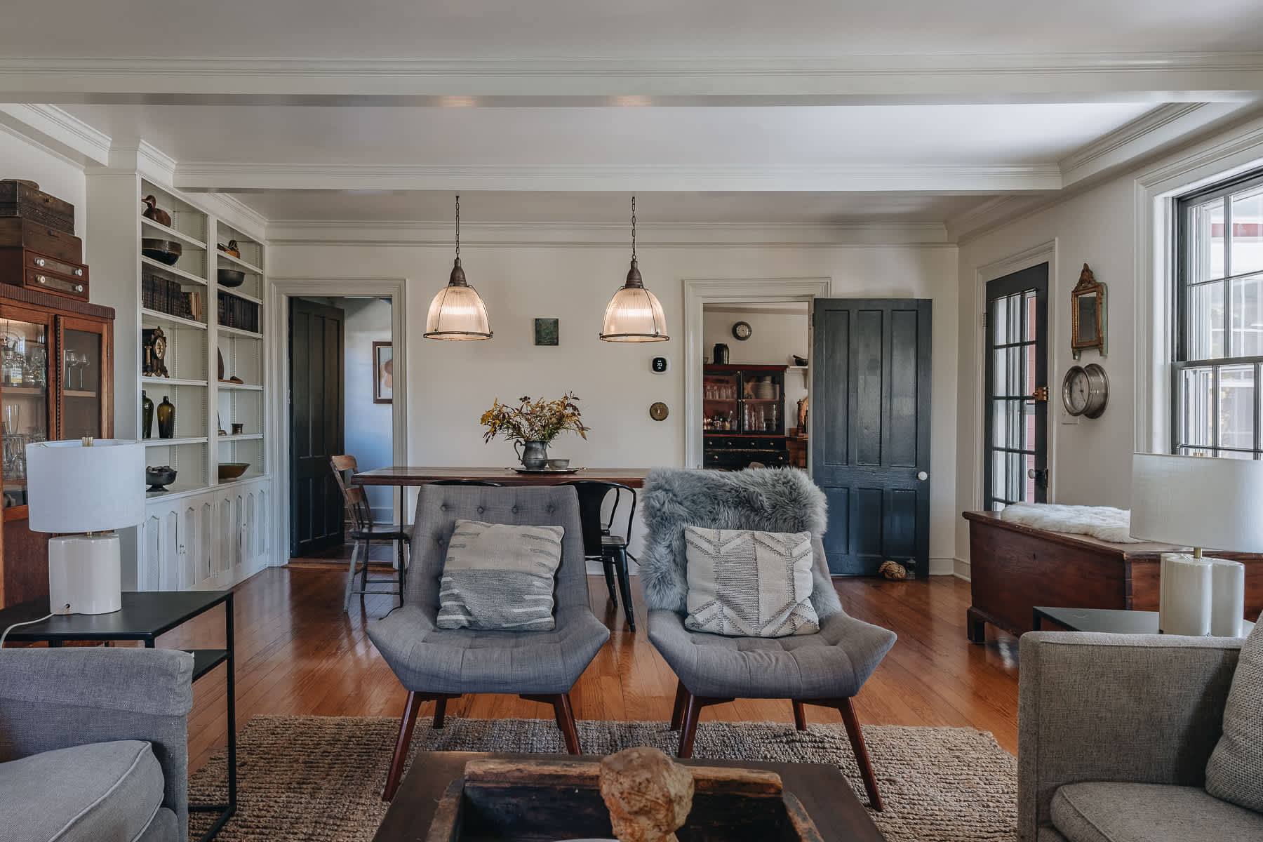 Open-concept living and dining area featuring a long wooden dining table with pendant lights, two gray armchairs with decorative pillows, and built-in shelving along the far wall.