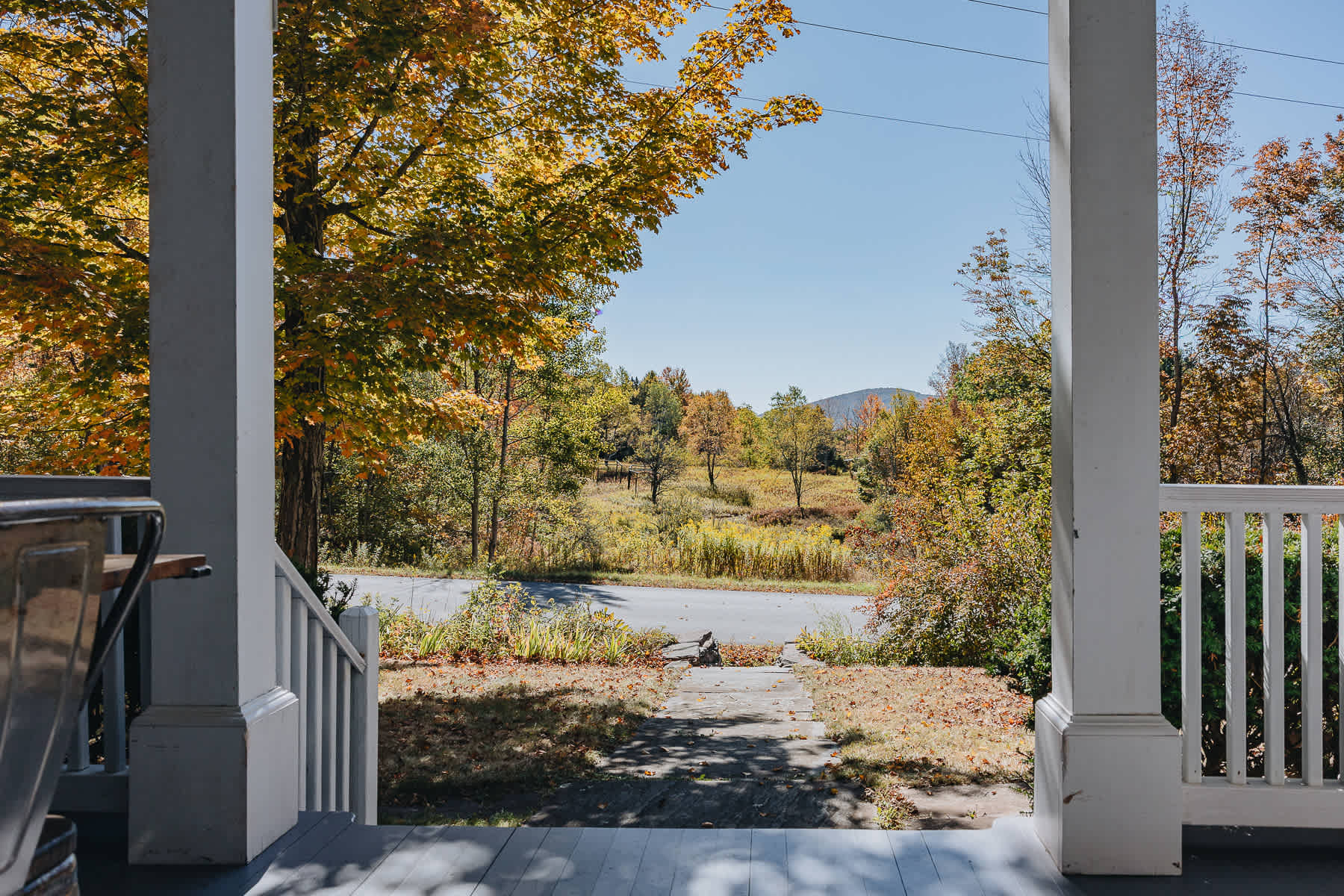 View from a white-railed porch overlooking a road and a vibrant autumn landscape with trees and distant hills under a bright sky.