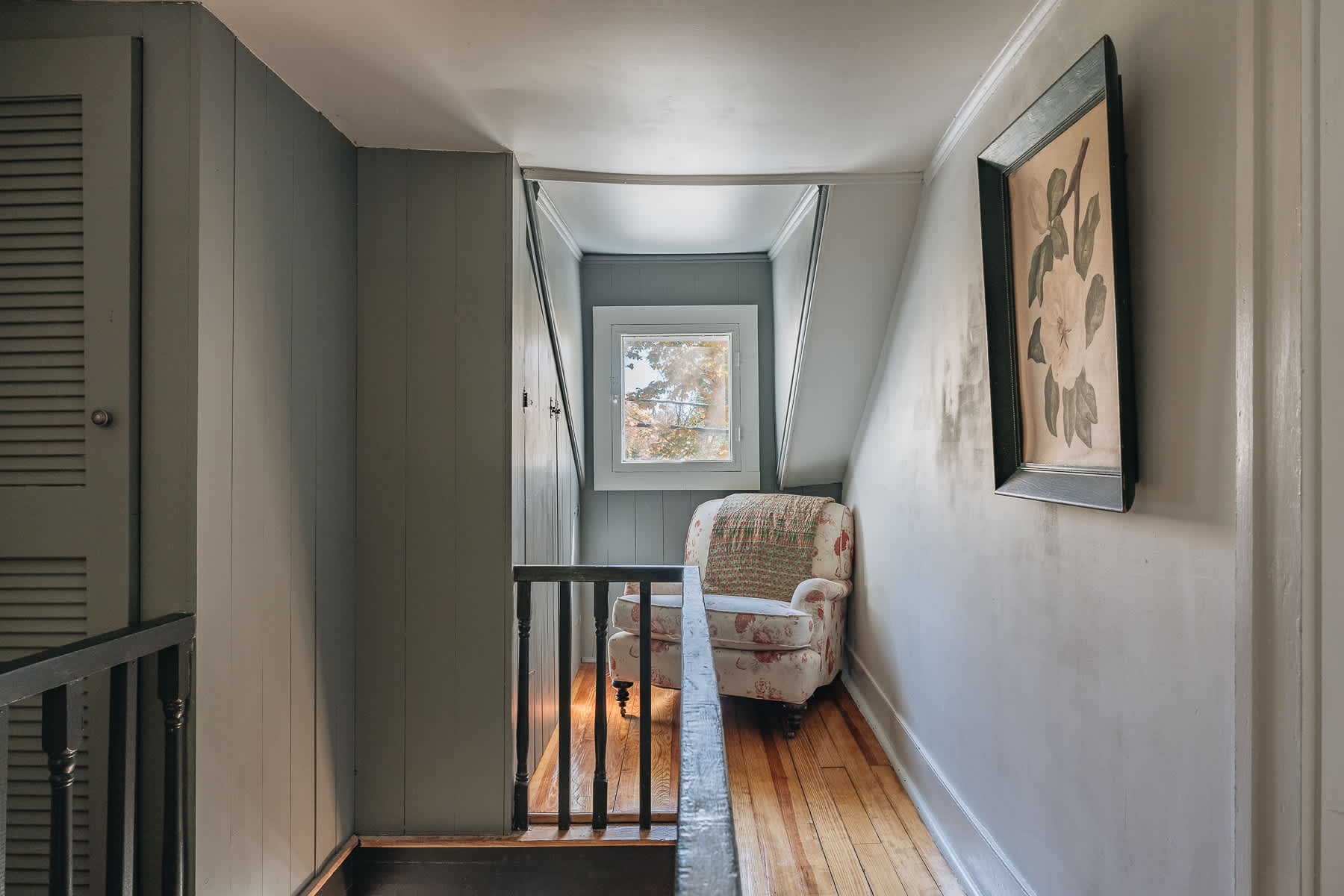 A narrow upstairs landing features sloped ceilings and light gray walls, with dark wood flooring and a black railing overlooking stairs. A cozy reading nook with an armchair and window is visible at the end of the hall.