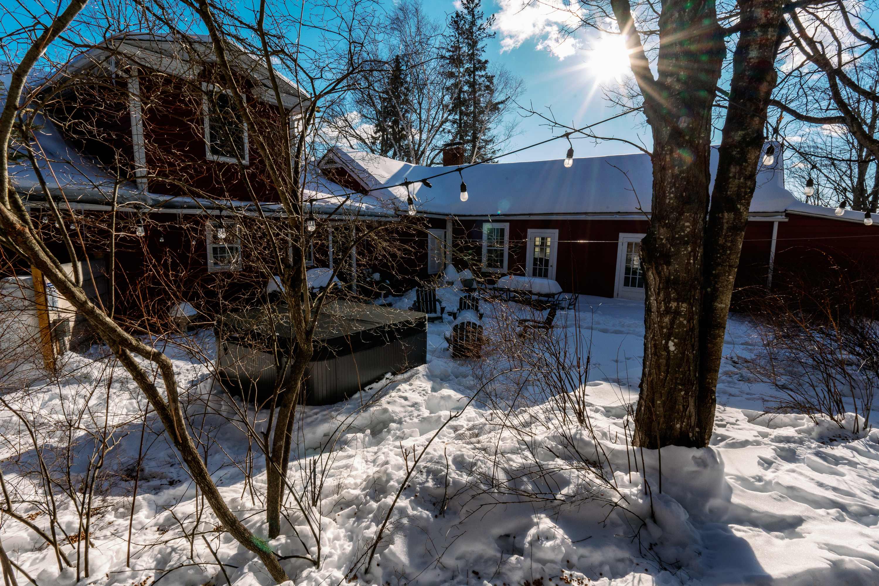 The back of a red farmhouse with white trim is covered in snow, featuring a dark hot tub, outdoor seating, and string lights draped between trees. Bright sunlight streams through the bare branches, creating a winter wonderland scene.