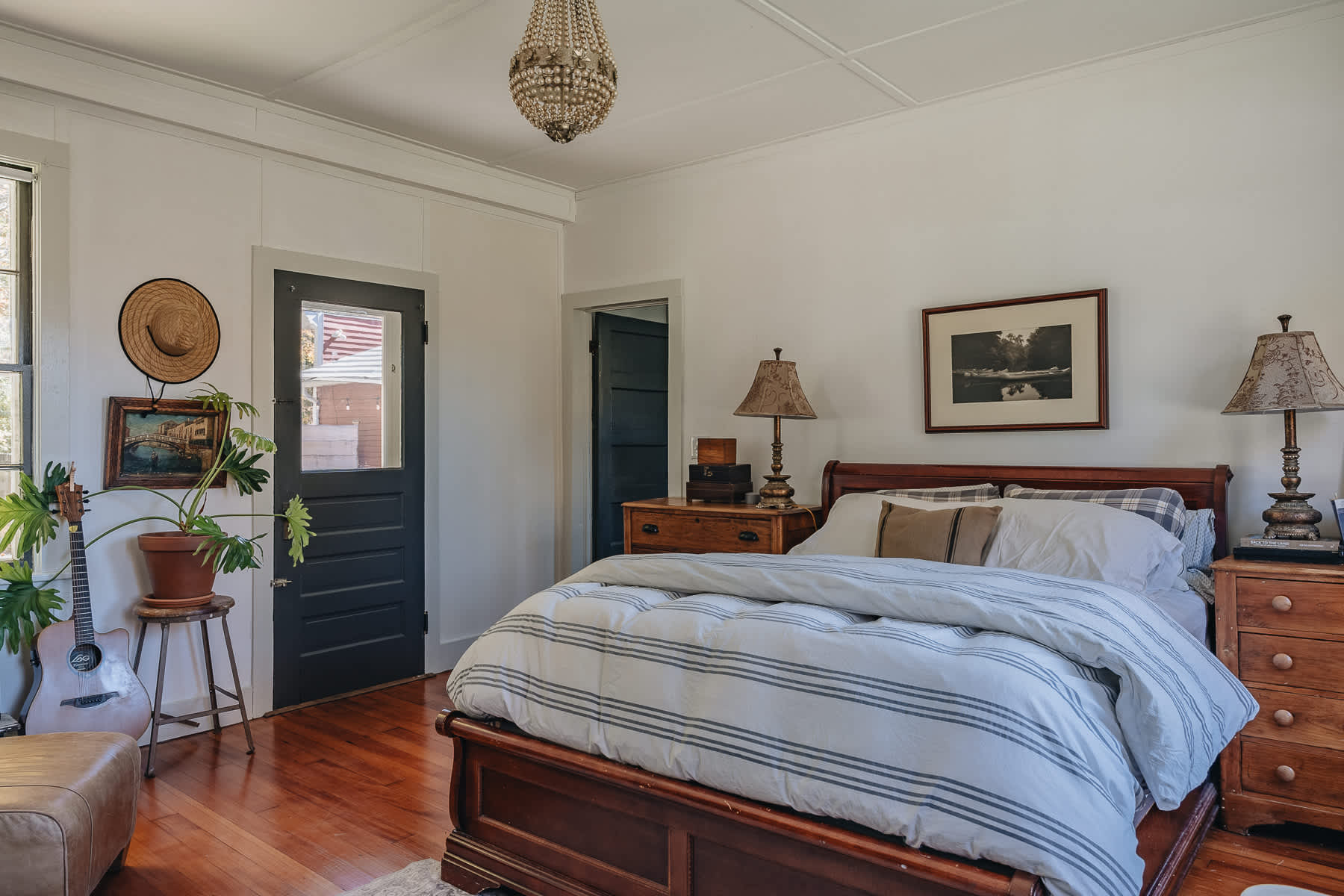 Cozy bedroom featuring a dark wooden sleigh bed with striped bedding, a wooden dresser with a lamp, and a dark paneled door leading outside. A guitar and plant are in the corner.
