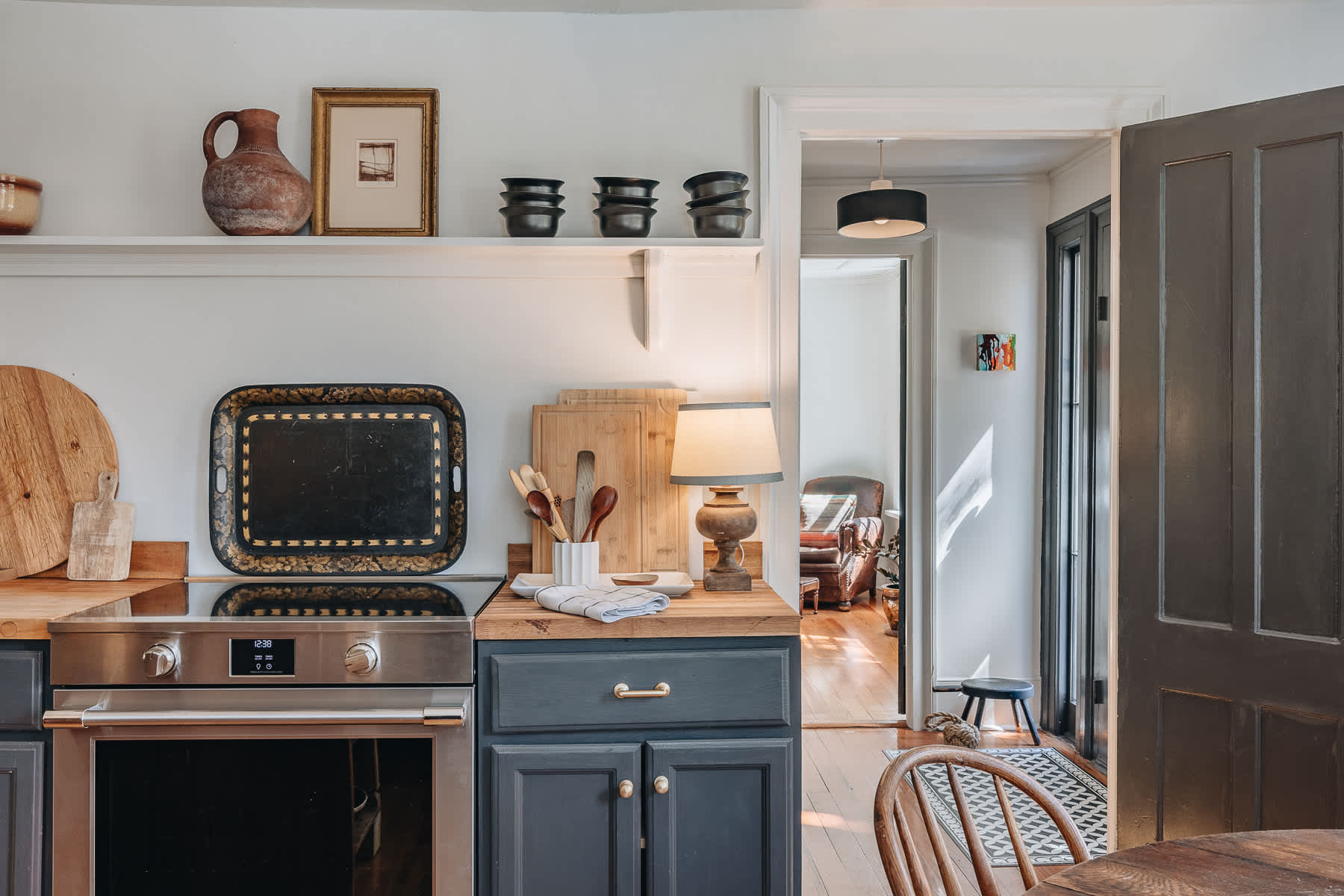 Rustic kitchen corner with a built-in oven, dark cabinetry, butcher block countertops, and open shelving displaying pottery. A dark wooden door is visible on the right.