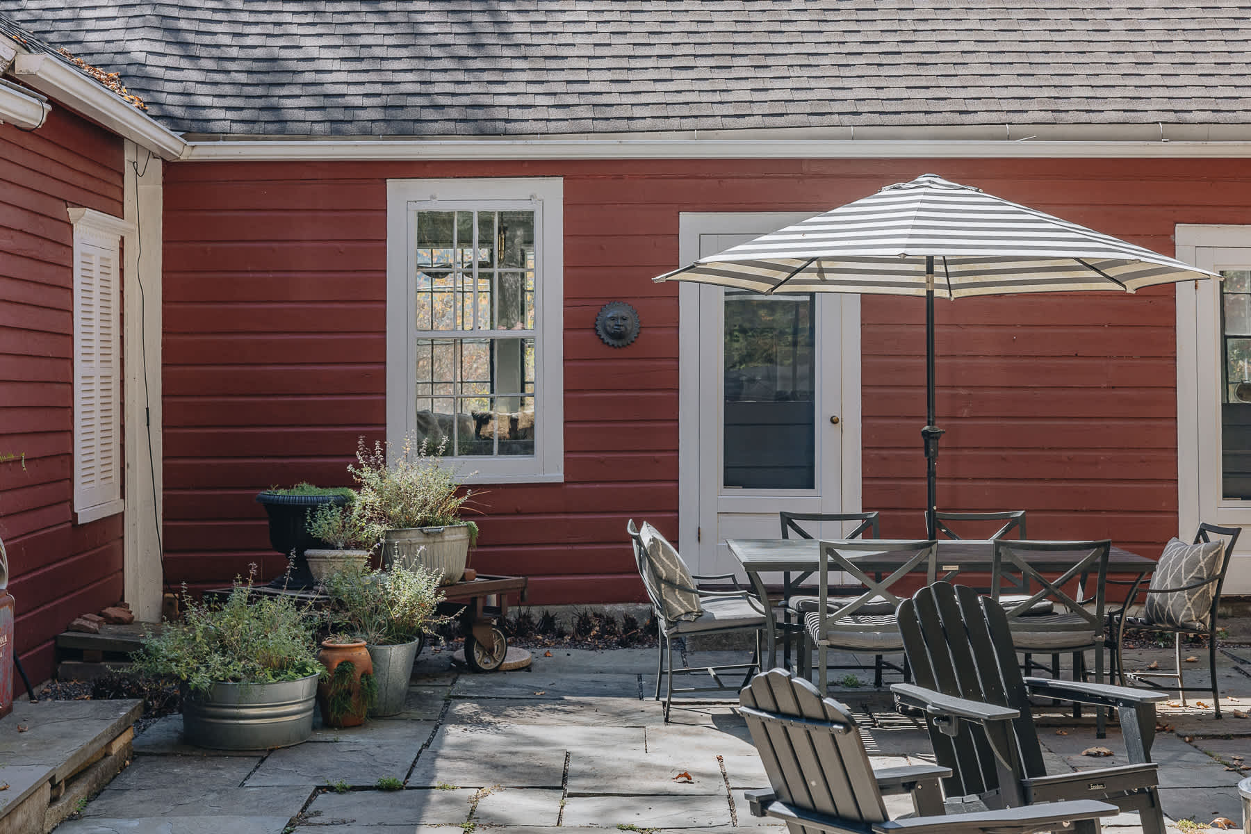 A stone patio features an outdoor dining set with a striped umbrella, surrounded by potted plants and the red exterior of the house. Adirondack chairs are also visible, suggesting a relaxed outdoor living space.