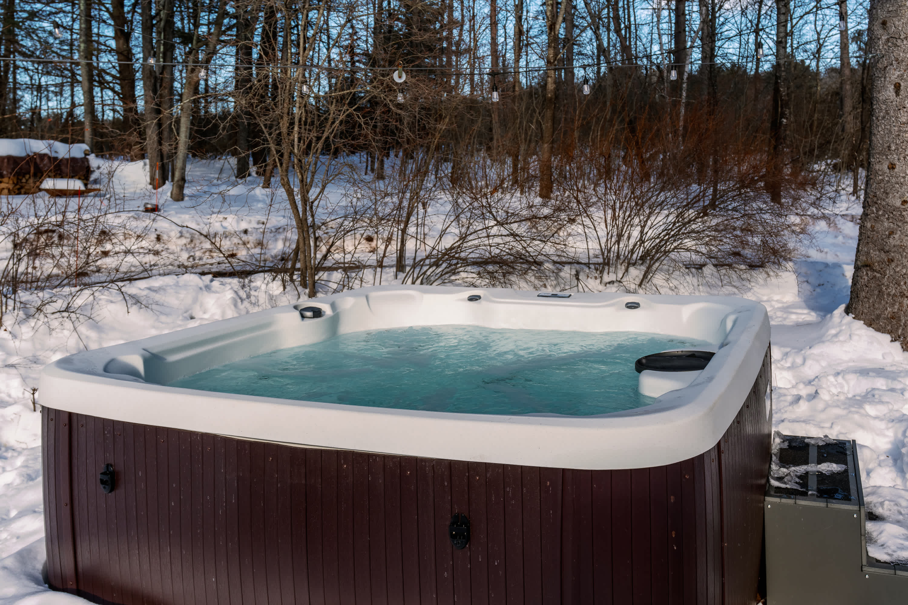 A wide shot of a hot tub in a snowy, wooded backyard, with stacked firewood visible in the distance. The scene highlights the hot tub as a winter amenity amidst a natural setting.